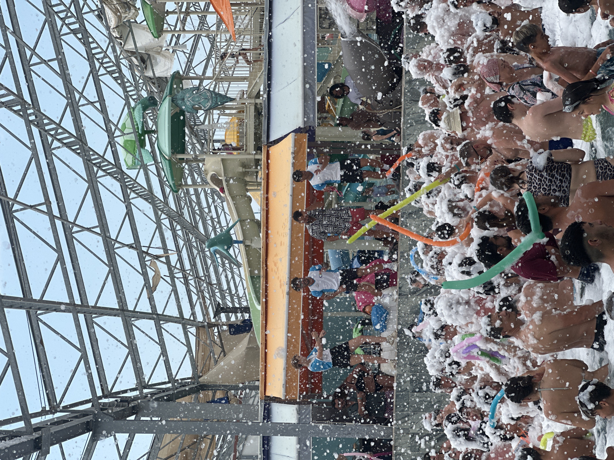 This image depicts a lively foam party taking place at a water park, likely in Kemer, Turkey, judging by the architecture and style which is common in the region's resorts. The scene is filled with people, many of them in swimwear, immersed in a large pool of white foam. The foam is so dense that it covers most of the individuals, creating a playful and somewhat chaotic atmosphere.
In the background, elements of a water park are visible, including colorful slides and structures that suggest water attractions. A metal truss roof system covers the area, with some sunlight filtering through, indicating it's an outdoor or semi-outdoor venue. The overall impression is one of summer fun and celebration, with participants enjoying the novelty of the foam. There are also inflatable balloons in various bright colors scattered amongst the crowd, adding to the festive mood.