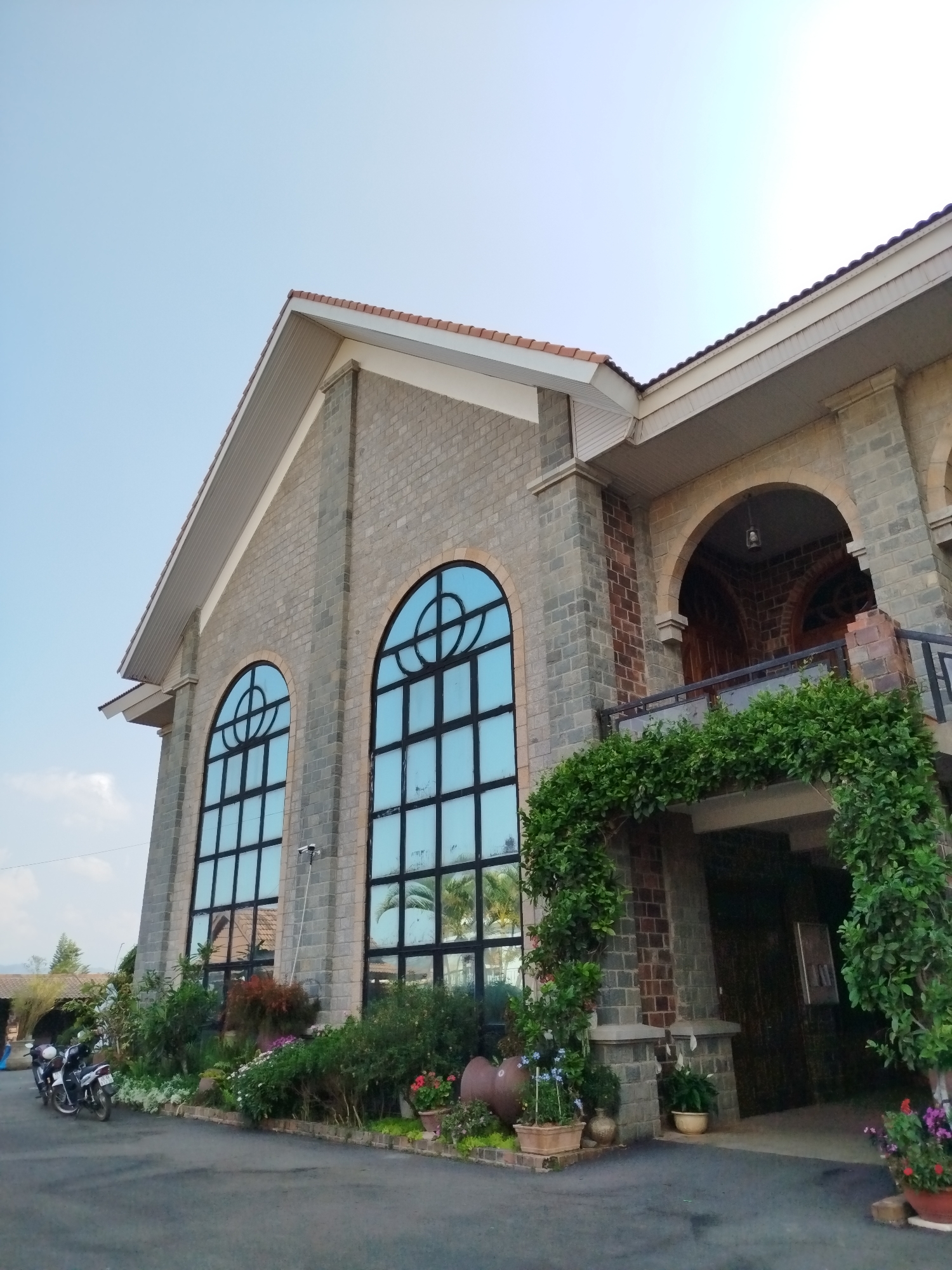 The image captures the exterior of a large, stone-clad building, possibly a church or a public institution, situated in Nam Hiệp 1, Vietnam. The scene is set outdoors under a clear, bright blue sky, suggesting daytime with soft, ambient lighting.
The building features a distinctive architectural style with a gabled, terracotta-tiled roof and an exterior constructed from rough-hewn, light grey stone blocks. The prominent facade showcases two imposing, arched windows with dark frames and reflective, light blue glass, which mirror parts of the sky and surrounding greenery. Above the main entrance area on the right, there is an upper-level balcony or loggia with arched openings and an ornate railing. Lush, green climbing vines cascade over a lower archway, framing an entrance.
Along the base of the building, a well-maintained garden bed with various plants, flowers, and potted arrangements adds natural vibrancy. Two motorcycles are parked on the left side of a paved driveway or forecourt in front of the building. Additional large, rustic ceramic pots are placed near the greenery. A long window cleaning tool is casually leaning against one of the large windows.
No people are visible, and no specific activity or event is taking place, giving the scene a serene and tranquil atmosphere. There is no discernible text visible anywhere in the image. The overall impression is one of a well-kept, stately structure nestled within a verdant setting.