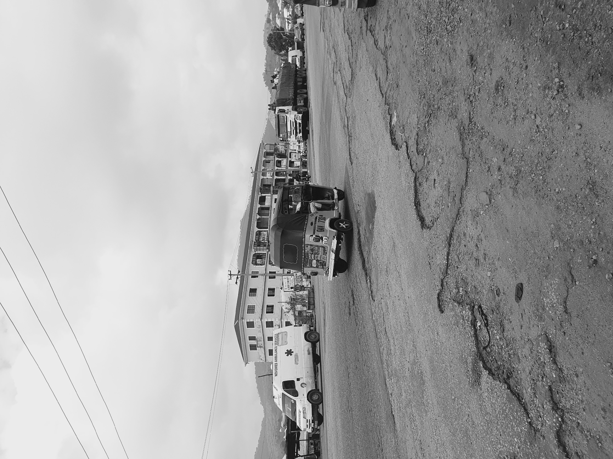 The image depicts a street scene in Lokoja, Nigeria. A road stretches from the foreground to the background. The road is heavily degraded with potholes and uneven surfaces. An ambulance is parked near a building, identified by the text on the side as belonging to "NovoMed Ambulance Services". A three-wheeled auto-rickshaw vehicle is also present, followed by a few other vehicles of different size. The background shows other buildings and vehicles, and in the distance, an undulating landscape is visible beneath an overcast sky. Power lines run across the top left corner of the image. There are no visible people in the image.