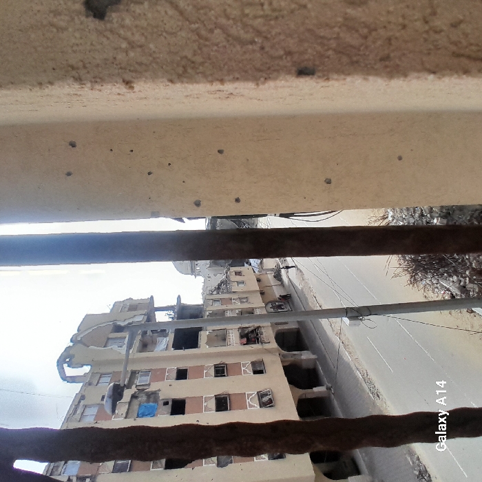A view through rusty bars reveals a damaged building across a street. The structure exhibits signs of significant decay and demolition, with missing walls and exposed interiors. The building appears to be in a state of ruin. A road runs beside the building, and utility poles are visible. The image is framed by concrete and bars, suggesting the vantage point is from another building. The scene depicts destruction and decay.