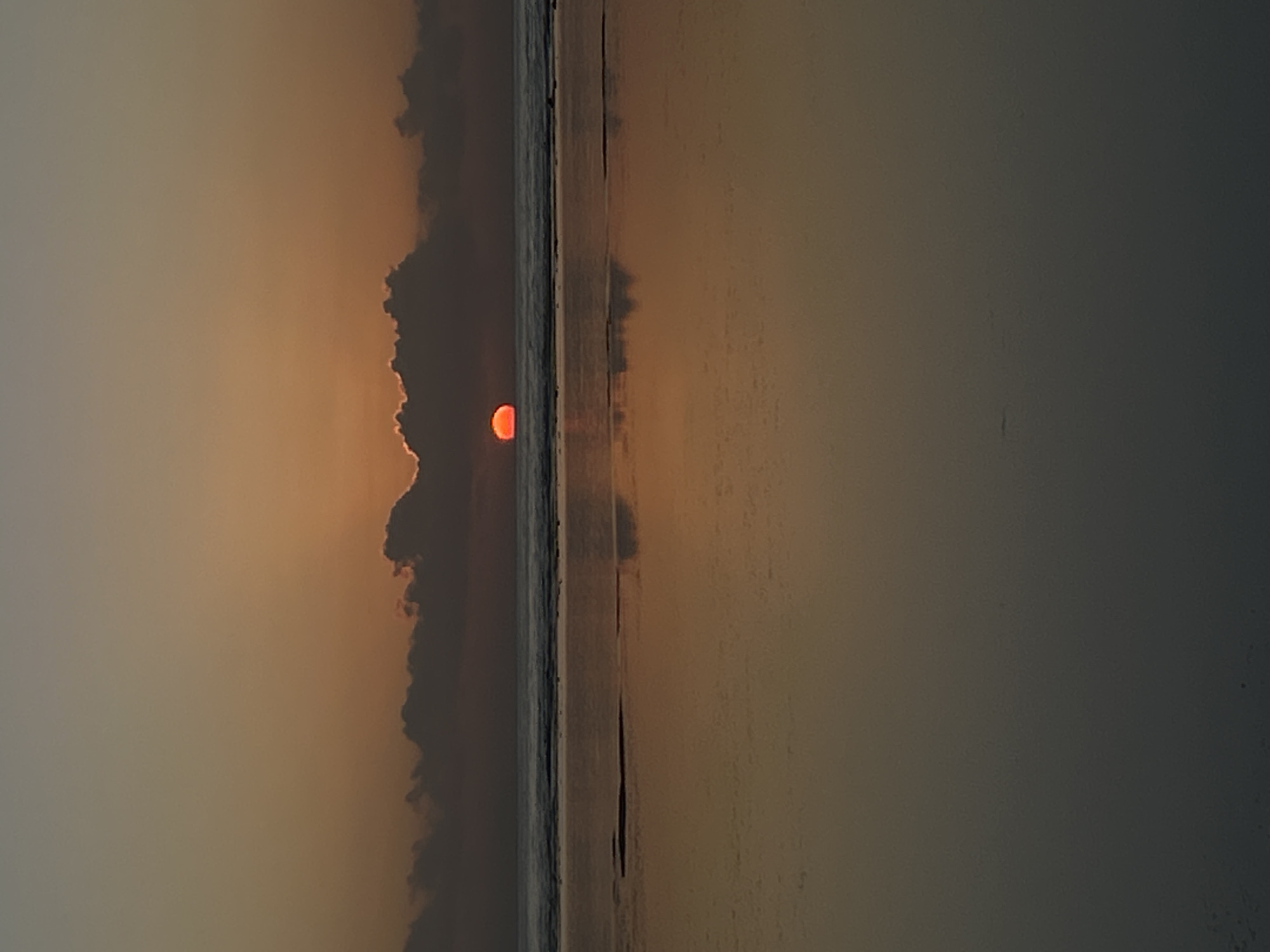 This is a photograph taken during sunset at a beach in Denpasar, Indonesia. The sun, a vibrant orange orb, is partially obscured by dark clouds, creating a dramatic and moody atmosphere. The sun's glow illuminates the sky with warm hues of orange and yellow, transitioning into a dusky gray above.
The scene depicts a body of water, likely the ocean, with gentle waves lapping at the shore. A thin strip of dark sand or land separates the water from the clouds and sky. The reflection of the sunset is visible on the calm surface of the water, adding depth and visual interest to the image. The overall impression is one of tranquility and the raw beauty of nature at dusk. There are no visible people or objects that are prominently featured. The weather appears calm, with the clouds adding a dramatic element to the otherwise serene sky. The time of day is clearly sunset, given the color of the sun and sky.