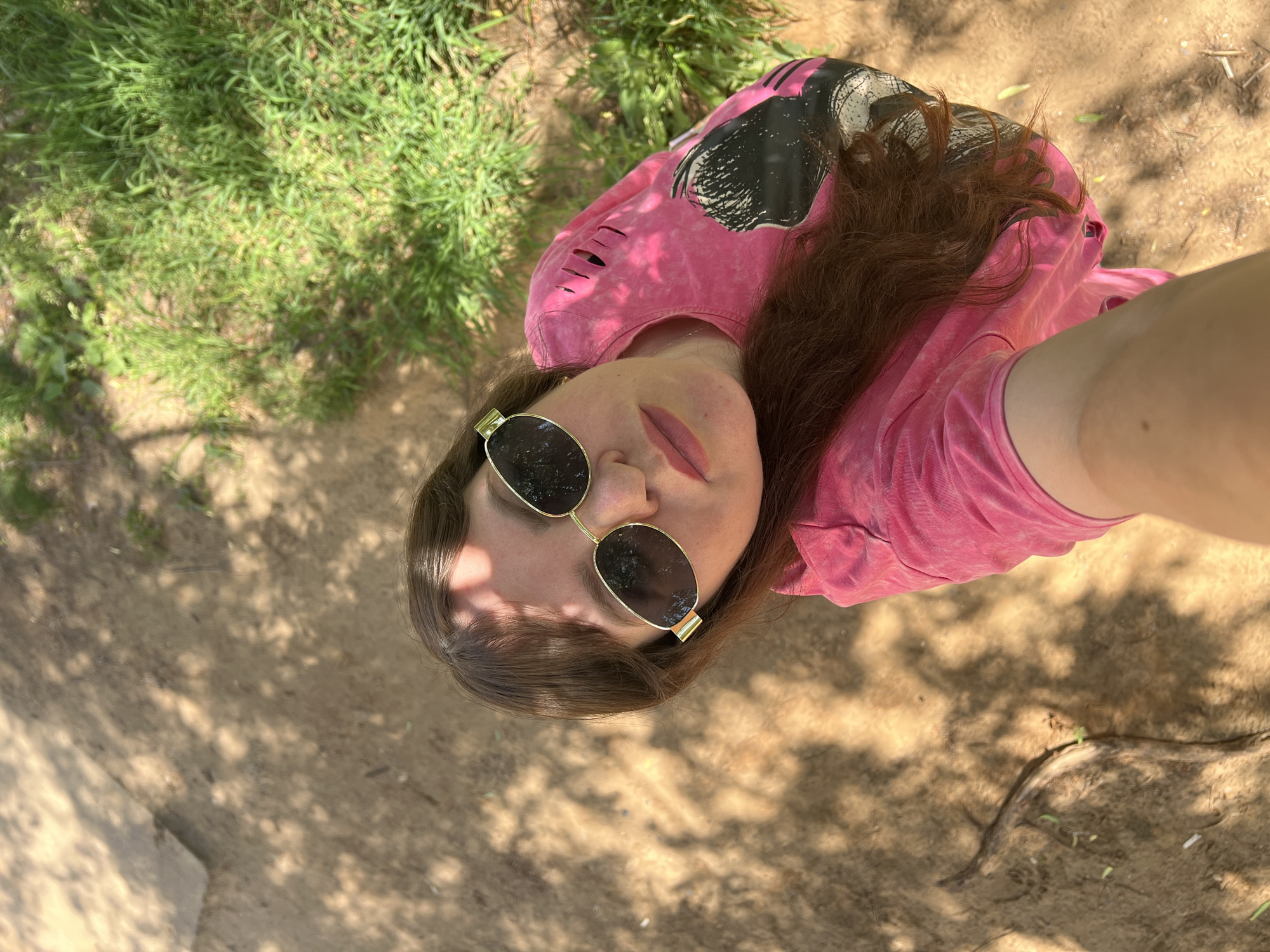A woman is taking a selfie outdoors. She is wearing a pink t-shirt with rips on the shoulder and a graphic design on the side. She has on gold-rimmed sunglasses and her brown hair is long and wavy. She is looking directly at the camera. The background is a mix of dirt ground with shadows from trees and green grass. The location is the city of Noviy, Russia.