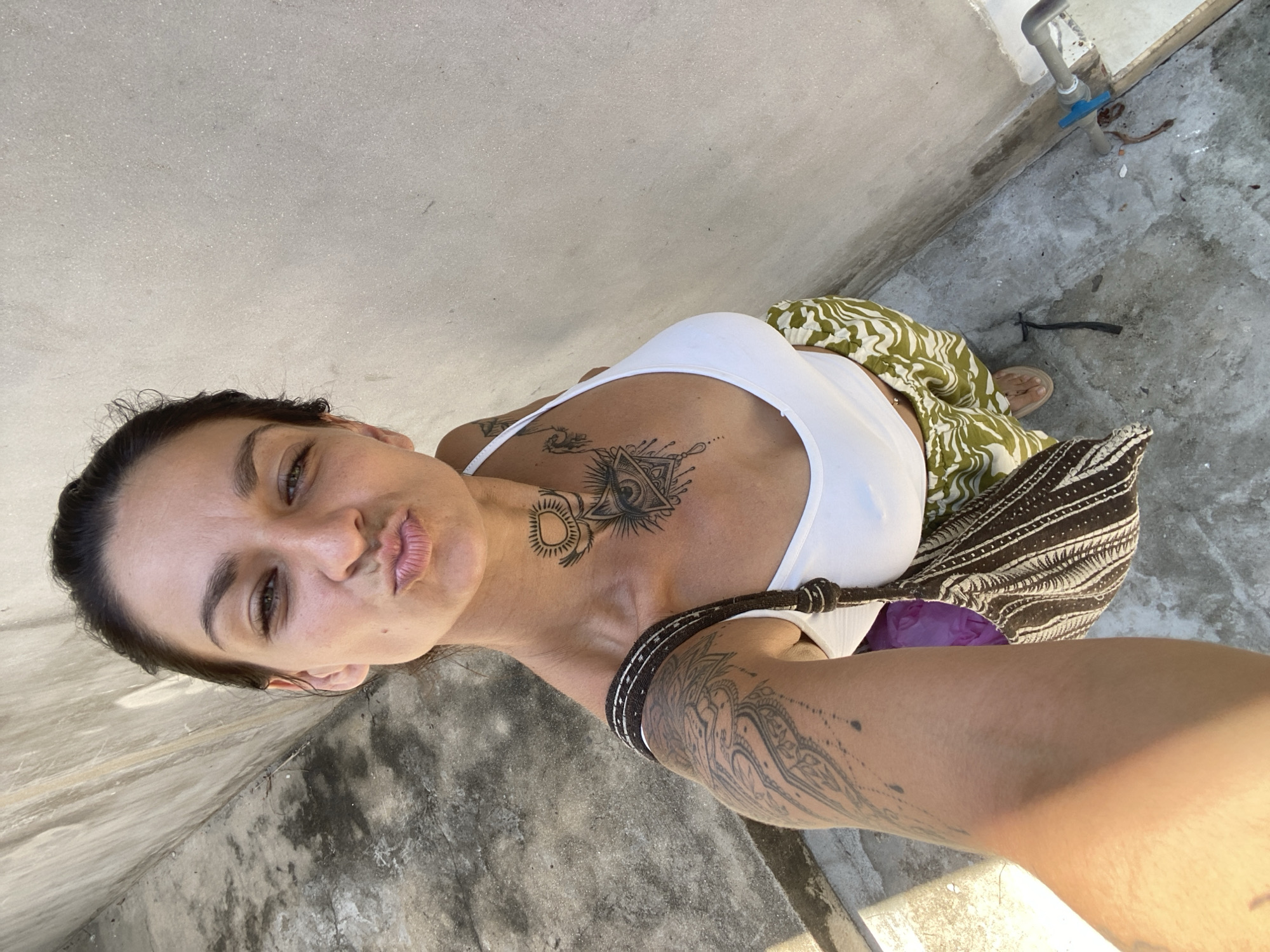 A young woman with dark hair, pulled back, faces the camera with her lips pursed in a kiss pose. She wears a white tank top, a patterned green and white skirt, and a patterned brown bag slung over her shoulder. She has numerous tattoos on her chest, neck, and left arm. She is standing on a concrete surface next to a textured, light-colored wall, with a pipe and a small puddle on the ground to her right. The scene appears to be an outdoor area, possibly a courtyard or alley, in Mũi Né, Vietnam, suggested by the architectural style and outdoor lighting.