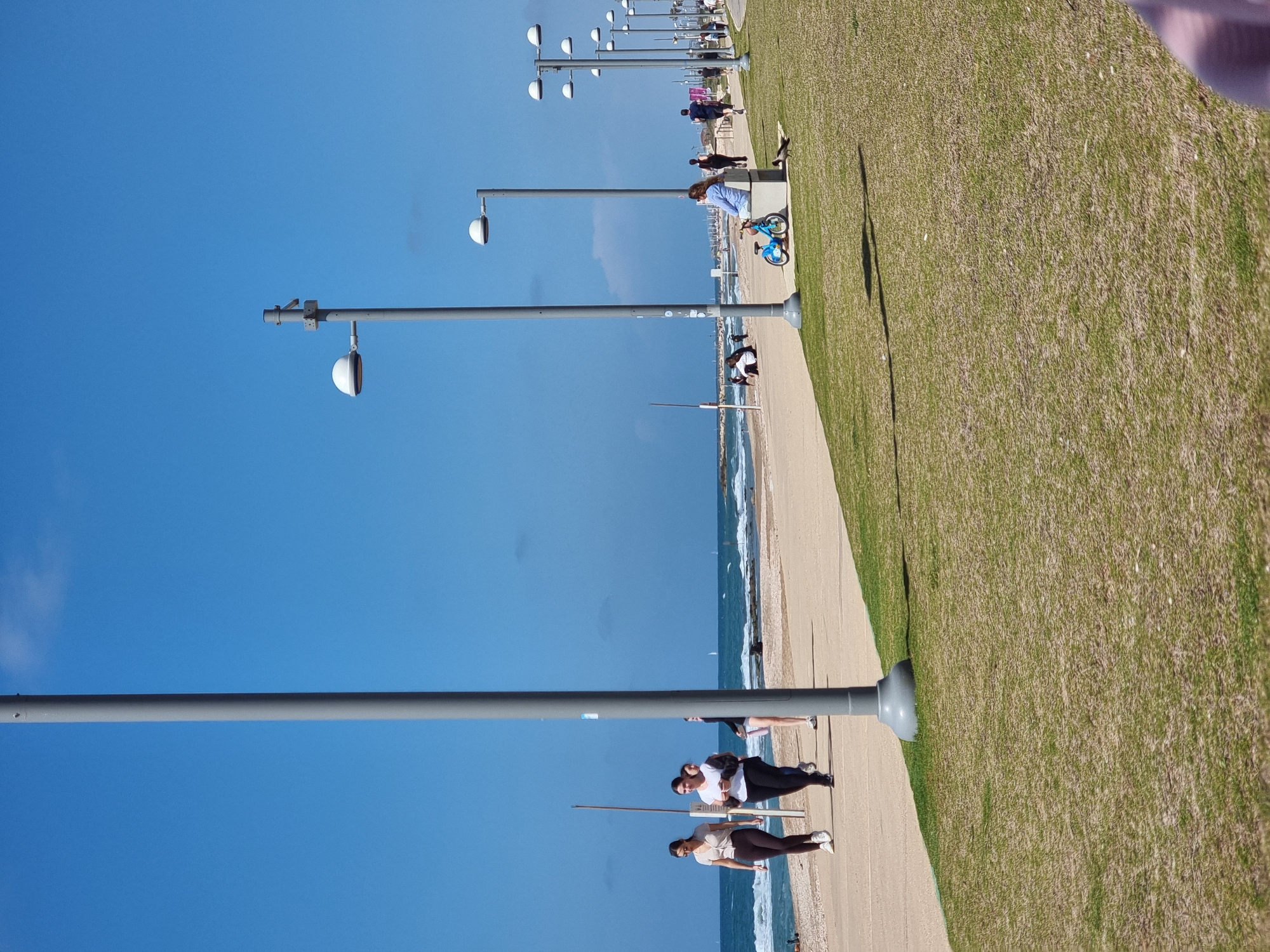 The image shows a street scene at a beach in Tel-Aviv, Israel. The photo is taken from an angle looking down the promenade. A row of equidistant lampposts lines the walkway. The sky is clear and blue. People are walking and biking along the paved walkway. The beach is to the left of the walkway with the ocean visible. A grass area is to the right of the walkway.
