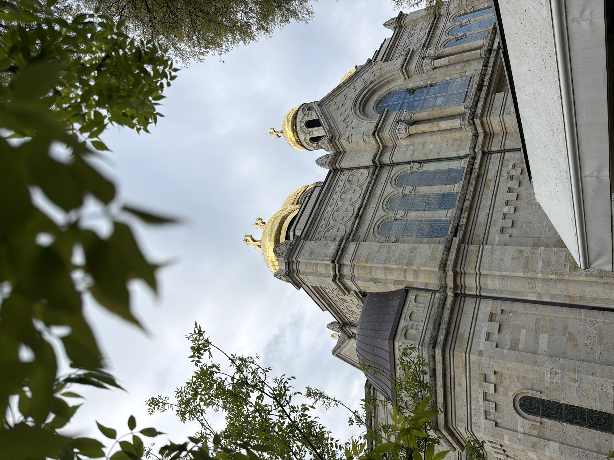 The image shows the Dormition of the Mother of God Cathedral in Varna, Bulgaria, from a low-angle perspective. The cathedral's façade is light-colored stone with architectural details such as arches, columns, and intricate carvings. Its golden domes are visible against a cloudy sky. Tree branches with green leaves frame the view on the left and partially at the top and bottom, adding a natural element to the urban scene. Part of a building, possibly an overhang, is visible on the right.
