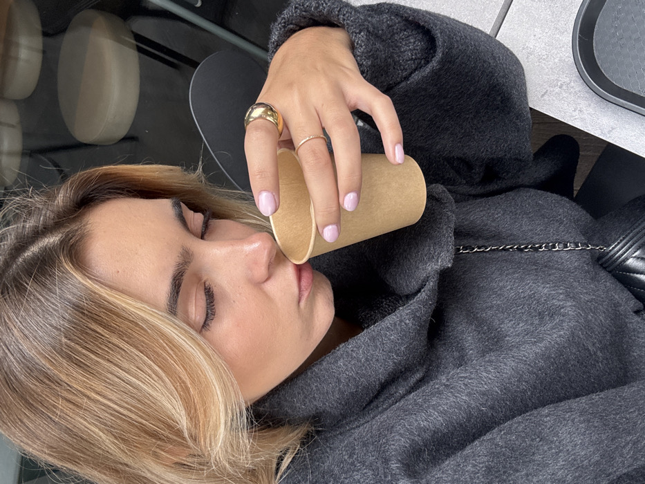 A young adult female is depicted in a close-up, high-angle shot, bringing a plain, light brown paper cup to her lips. Her eyes are closed, and her head is tilted slightly downward, indicating focus on the object or its contents. She has blonde hair with darker roots, parted on the side, and well-defined dark eyebrows. Her skin is light-toned, with visible cosmetic enhancement around the cheeks. She is wearing a dark gray, textured, cowl-neck garment, possibly a sweater or coat. Her left hand, adorned with light pink nail polish, holds the cup. A large, polished gold-colored domed ring is on her left ring finger, and a thin, gold-colored band is on her left middle finger. A black quilted bag with a silver-colored chain strap is partially visible below her left arm. The background includes reflections of circular light-colored objects, possibly stools or tabletops, in a dark, reflective surface, along with a dark chair element. A light gray, textured tabletop and a black rectangular tray or plate are visible in the lower right foreground. The scene is situated in Vaulx-en-Velin, France.