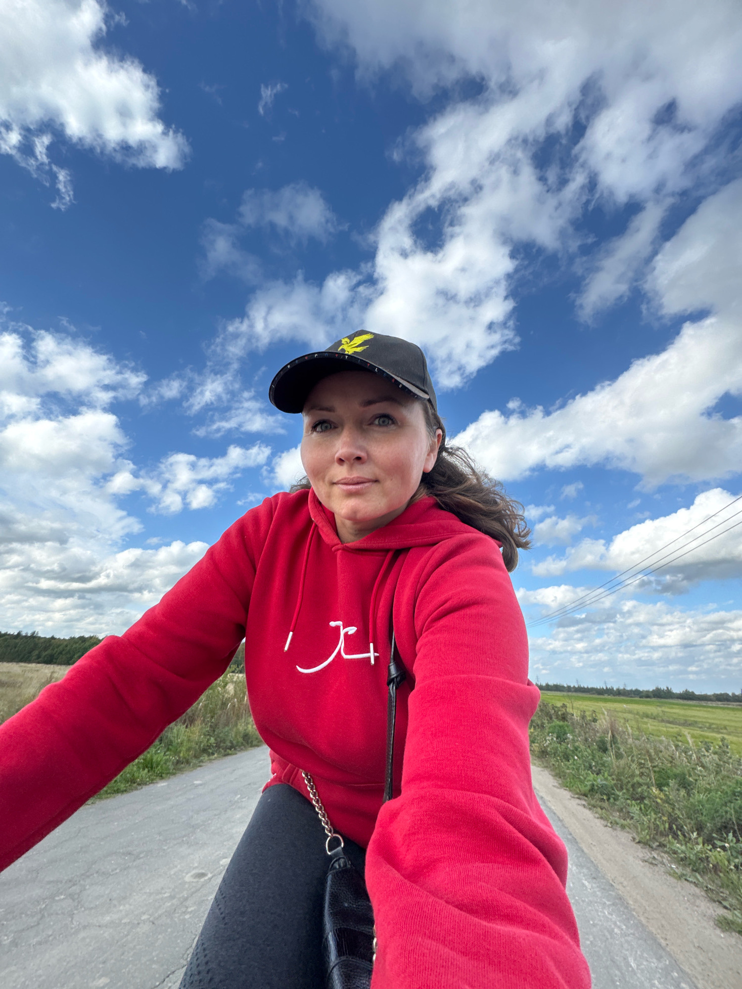 A woman wearing a red hoodie and a black baseball cap is shown from the chest up, riding a bicycle on a paved road. The road is bordered by tall green grass and a distant forest. The sky above is a bright blue with scattered white clouds. The woman is looking slightly to her left, with a neutral expression. A black purse with a gold chain strap is visible hanging from her shoulder. The overall scene suggests an outdoor activity in a rural or suburban setting. The location is identified as Innolovo, Russia.