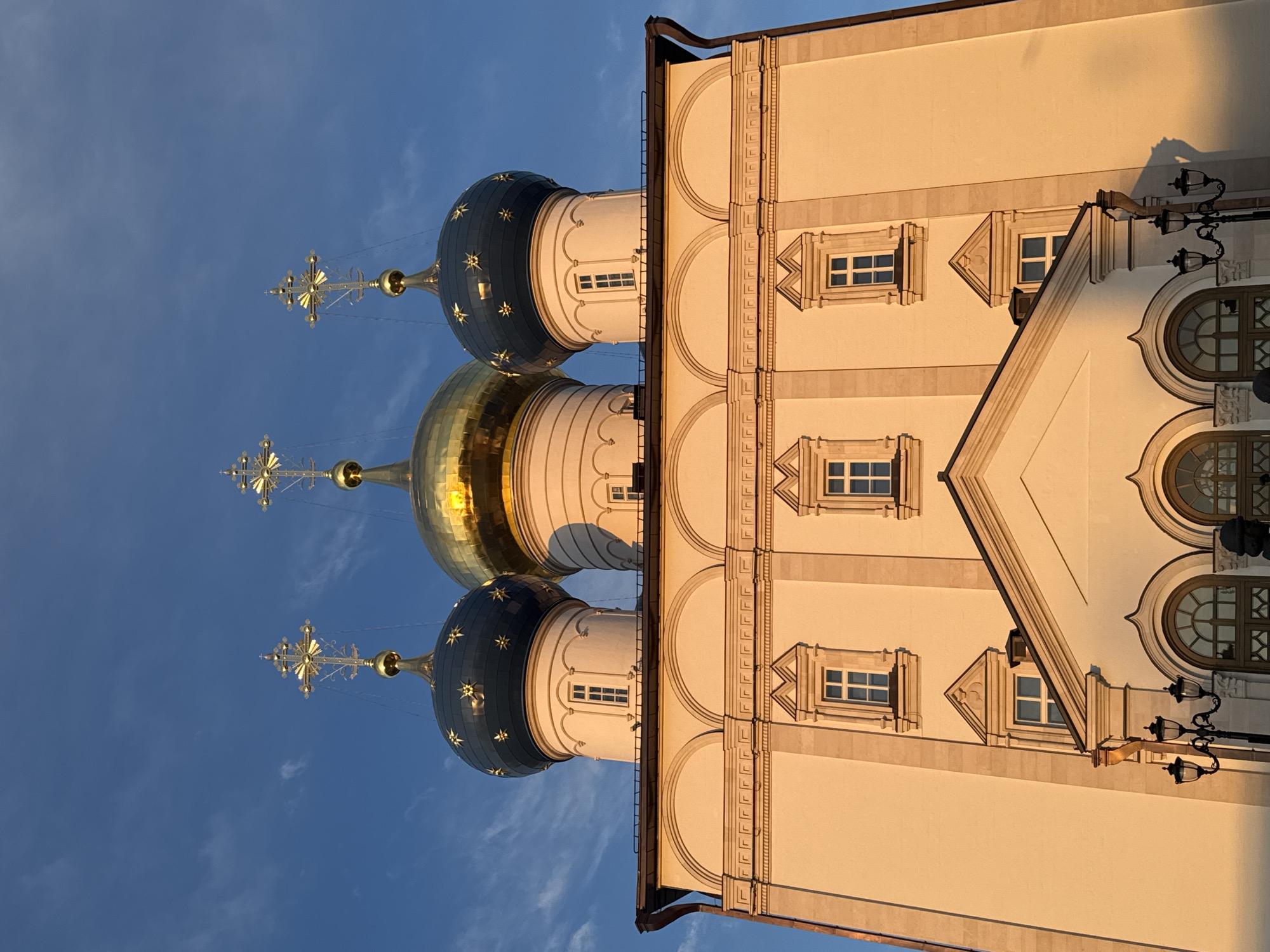 The image captures a low-angle perspective of the Church of the White Trinity in Tver, Russia, against a clear blue sky. The church's architectural features are prominent, including its three onion-shaped domes, two of which are dark blue with golden stars and one is entirely gold. Golden crosses atop spires rise from the domes.
The facade of the church is visible, exhibiting a cream-colored exterior with decorative architectural details around the windows and arches. The lighting suggests it is either sunrise or sunset, casting a warm glow on the building. A vintage-style lamppost is attached to the building on the right side. There are no people visible in the frame.