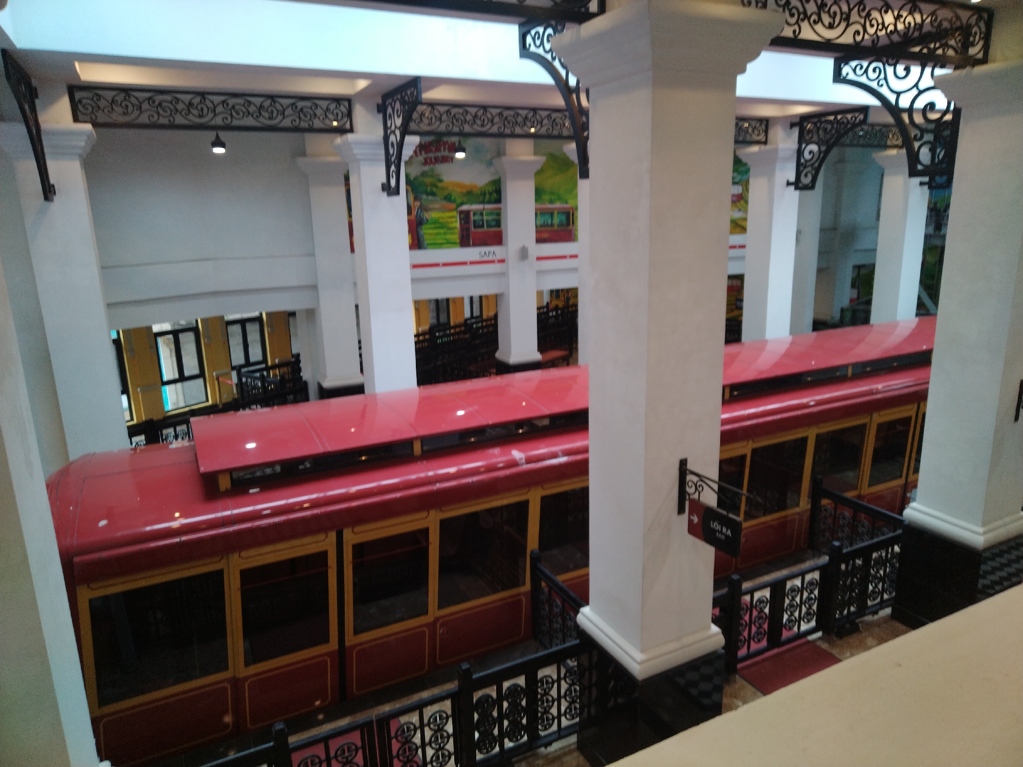 The image displays an indoor funicular station or a similar transport hub, characterized by a multi-level structure and bright illumination. Two parallel red train cars, featuring numerous rectangular windows with golden-brown frames, are prominently positioned within a deep, track-lined trench or pit, viewed from an elevated vantage point.
The architecture includes several robust white pillars that support the upper levels and roof. Ornate black wrought-iron railings with intricate scroll patterns line the platforms and stairways, and similar decorative ironwork adorns the ceiling cornices, adding an elegant touch to the industrial setting.
On the walls above the train tracks, colorful murals depict scenic mountainous landscapes, complete with green terraced fields and what appear to be funicular trains winding through them. The word "SAPA" is clearly visible in white letters on one of these murals, strongly indicating the location as Sa Pa.
A black and red directional sign is affixed to one of the white pillars. It features a right-pointing arrow and text that reads "LÔI RA" in larger white letters, with "EXIT" printed below in smaller font. No people are visible, and the trains appear stationary at what is likely a boarding or alighting platform. The scene suggests a well-maintained and aesthetically pleasing transportation facility, likely catering to tourism in Sa Pa Ward, Vietnam.