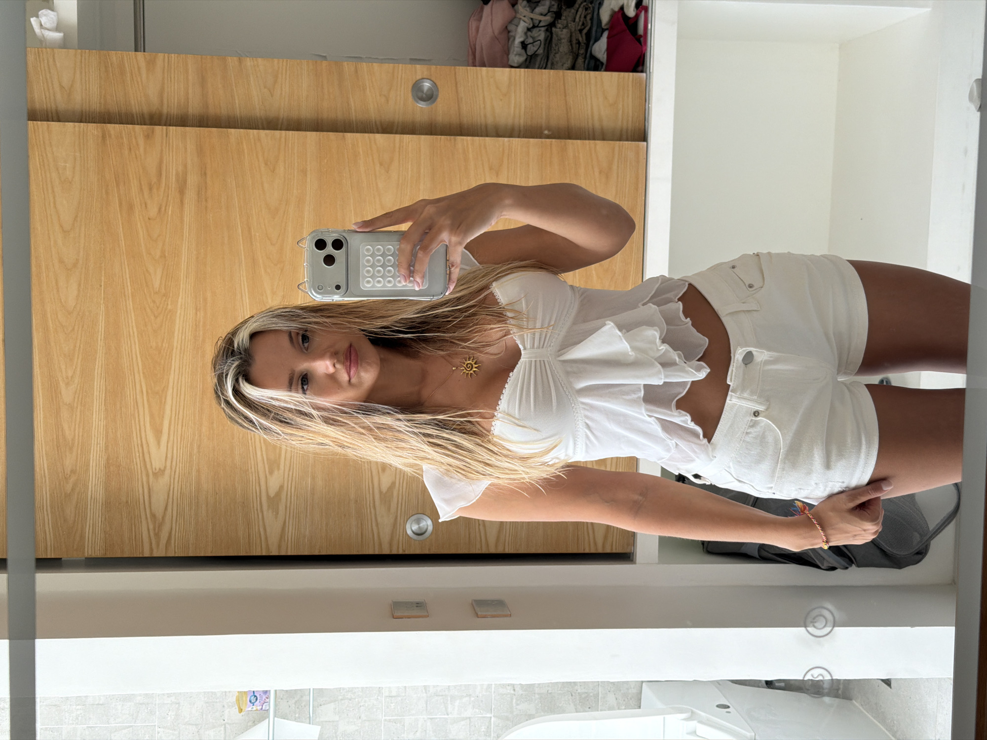 A young woman with wet blonde hair is taking a mirror selfie in a room. She is wearing a white ruffled crop top and white denim shorts. She has a gold sun pendant necklace and a colorful bracelet on her left wrist. She is holding a silver smartphone with a circular button pattern on the case. The background shows a wooden closet with sliding doors and shelves filled with clothing. To the left, a white toilet is visible. The scene is likely a bathroom or bedroom in Thep Krasatti, Thailand.