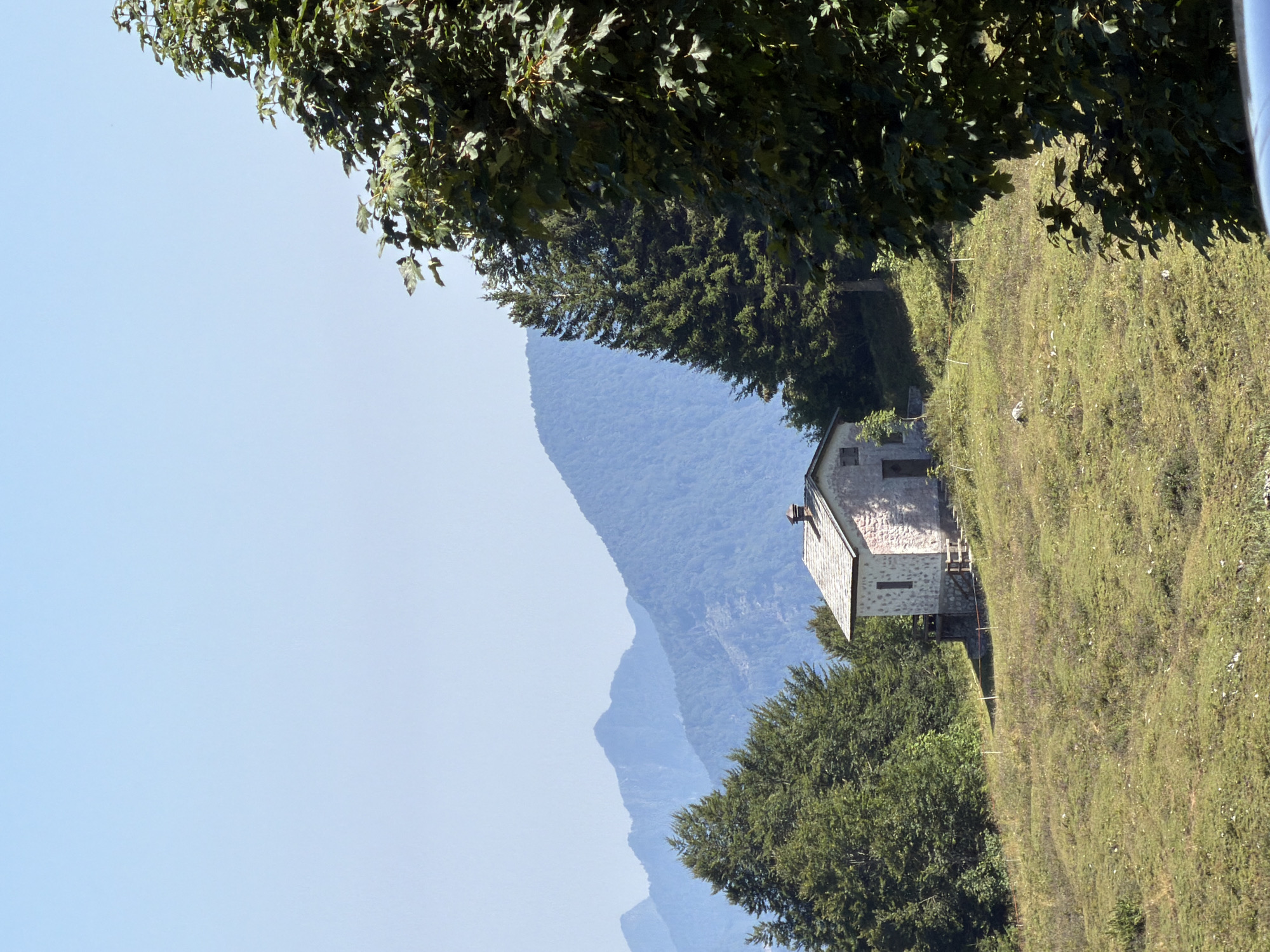 The image depicts a stone mountain hut situated on a grassy slope in Ampezzo, Italy. The hut has a gray, pitched roof and appears to be constructed from light-colored stones. It is surrounded by lush green trees, including a large, leafy tree in the foreground to the right. In the background, a series of progressively distant mountain ranges are visible against a clear blue sky. The terrain is characterized by rolling hills covered in grass and scattered vegetation. The overall scene suggests a remote, natural setting typical of the Italian Alps.