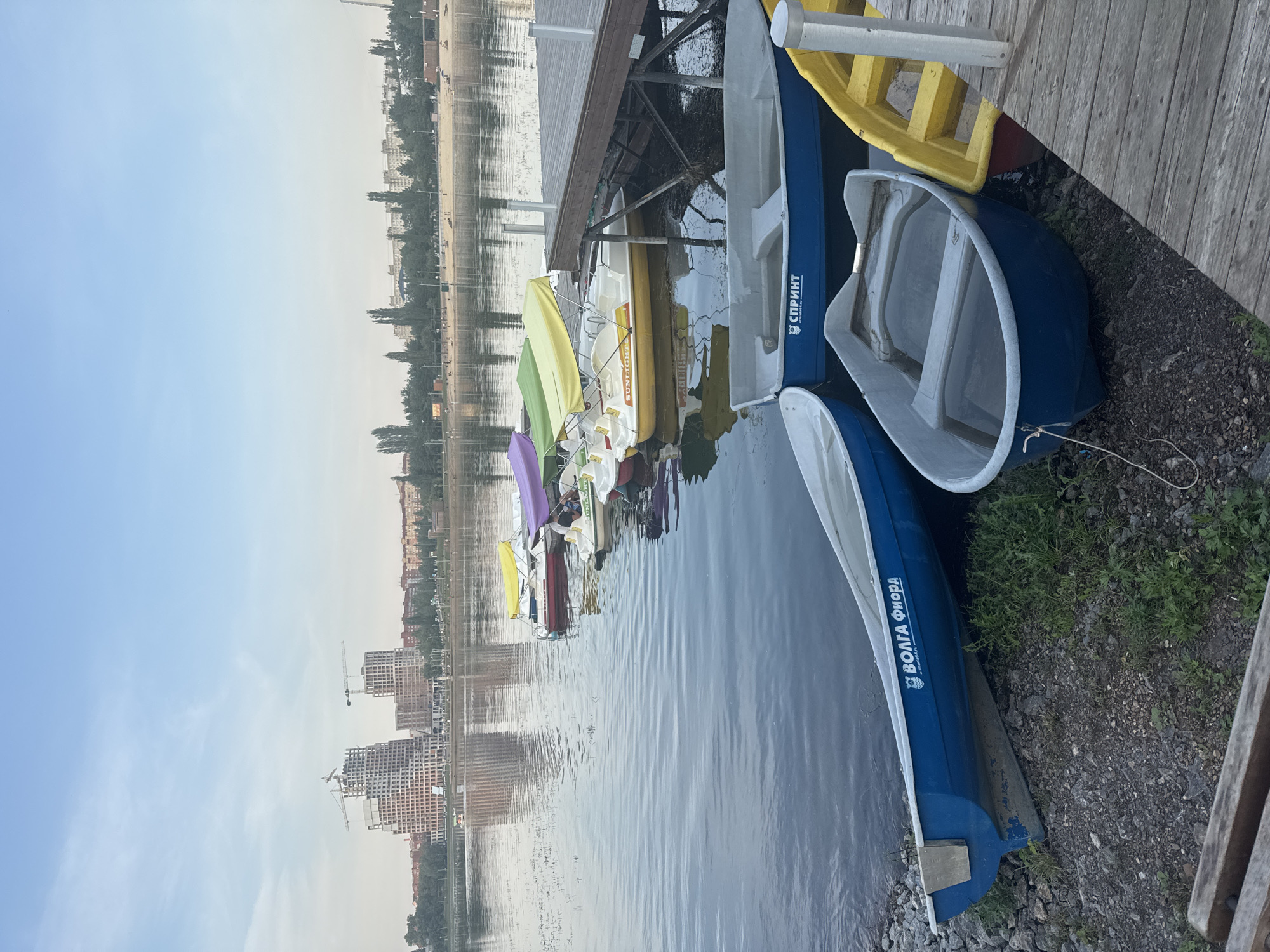 The image depicts a waterfront scene in Ufa, Russia, likely along the Belaya River. In the background, unfinished high-rise buildings under construction dominate the skyline, suggesting ongoing development in the city. A cluster of paddle boats with colorful awnings are moored near a wooden pier. Closer to the foreground, several rowboats are visible. One prominent blue rowboat has Cyrillic text visible on its side, reading "ВОЛГА ФИОРА". Another blue rowboat, partially submerged, sits on the rocky shore with a rope tied to it. A yellow paddleboat is also visible near the pier. The water is calm, reflecting the sky and the buildings. The shoreline is covered with gravel and sparse vegetation. The lighting suggests it is either early morning or late evening. There are no people clearly visible in the immediate foreground or interacting with the boats.