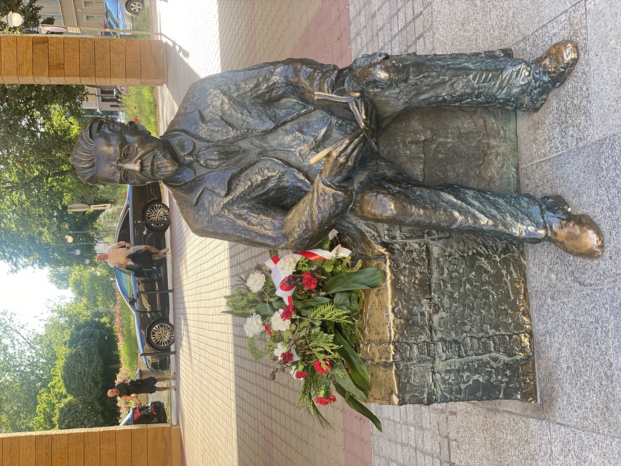 This is a full-body, eye-level shot of a bronze statue of a man seated on a stone block. The man is depicted wearing a suit, glasses, and a goatee. His hands are resting on a book. A floral arrangement is placed on the stone block to his left, featuring red and white carnations and greenery. The statue is located in front of a building with a textured, light brown facade and a pillar. In the background, two men are visible near cars. One man, wearing a black sleeveless shirt and shorts, is standing next to a black car. The other man, in a white t-shirt and shorts, is exiting another black car, holding a black backpack. The scene is set outdoors on a paved walkway under a sunny sky, with trees and other buildings in the distance. The statue is located in Łódź, Poland, specifically in front of the Faculty of Electrical Engineering, Automatic Control, Computer Science and Biomedical Engineering of the Lodz University of Technology, on ul. Sterlinga 16/18.