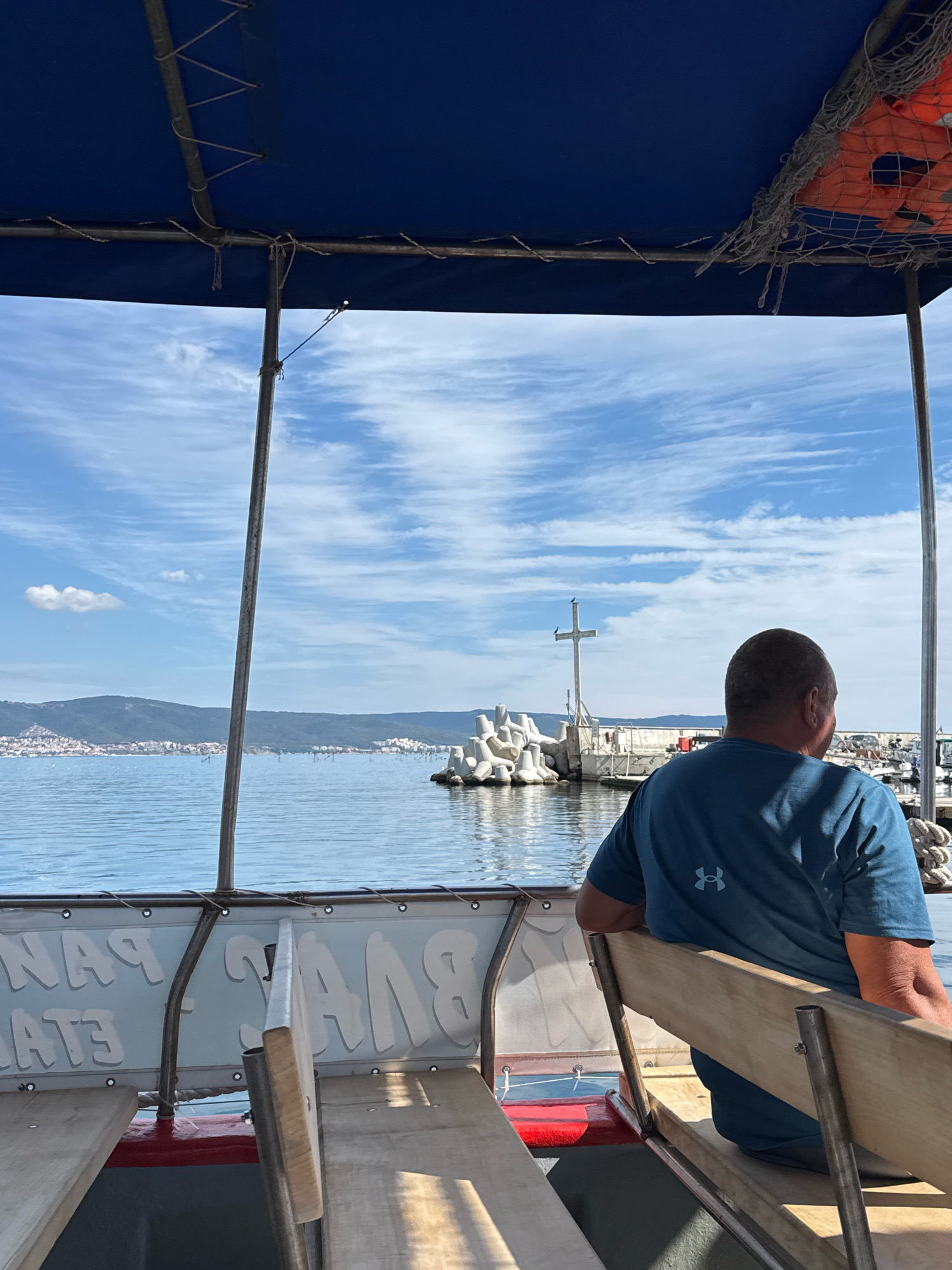 An outdoor, medium shot captures a man from the back, seated on a boat in the sea, facing away from the camera. He is wearing a dark blue t-shirt with an Under Armour logo visible on the back. The boat has wooden benches and a blue canopy overhead, with life vests visible in netting attached to the canopy. The man is looking towards a body of water where a town with white buildings is situated on a hilly coastline in the distance, identifiable as Nesebar, Bulgaria. A cross monument stands on a breakwater in the middle ground, with concrete structures and a harbor with several boats to the right. The sky is blue with scattered white clouds. The overall scene depicts a leisurely boat trip with a view of the coastal town.