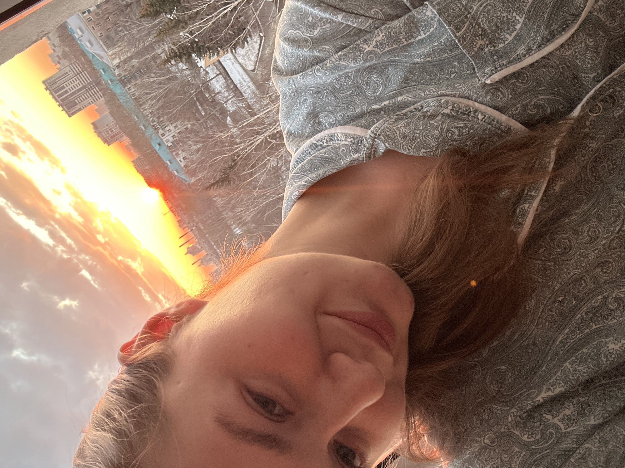 A selfie shot shows a person with fair skin, light brown hair, and wearing patterned pajamas. The person is positioned in front of a window with a view of a cityscape at sunset. The buildings are silhouetted against a bright orange and yellow sky, indicating either sunrise or sunset. There are trees visible, some with bare branches, suggesting a fall or winter season. The city is of Unknown, Unknown.