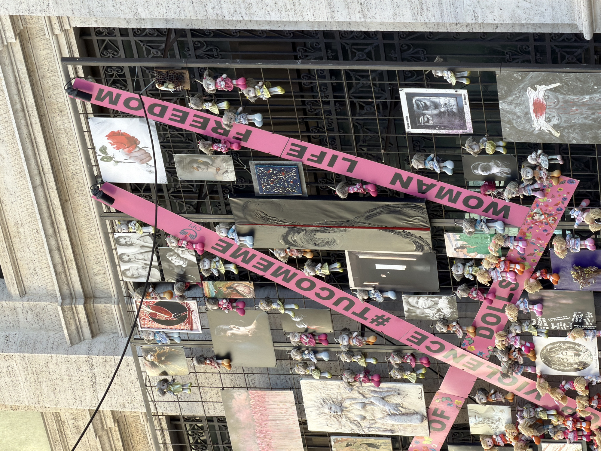 The image depicts an artistic installation in Genoa, Italy, attached to a building's exterior. The installation consists of a metal grid filled with various artworks (photographs, prints, drawings) and numerous small dolls. Two pink banners intersect diagonally across the grid, displaying the messages "WOMAN LIFE FREEDOM" on one and "#TUCOME ME NO MORE VIOLENCE OF DOLLS" on the other. The building's facade is visible on the left side of the frame, showing a stone texture. The scene is captured from a low angle, looking upwards.