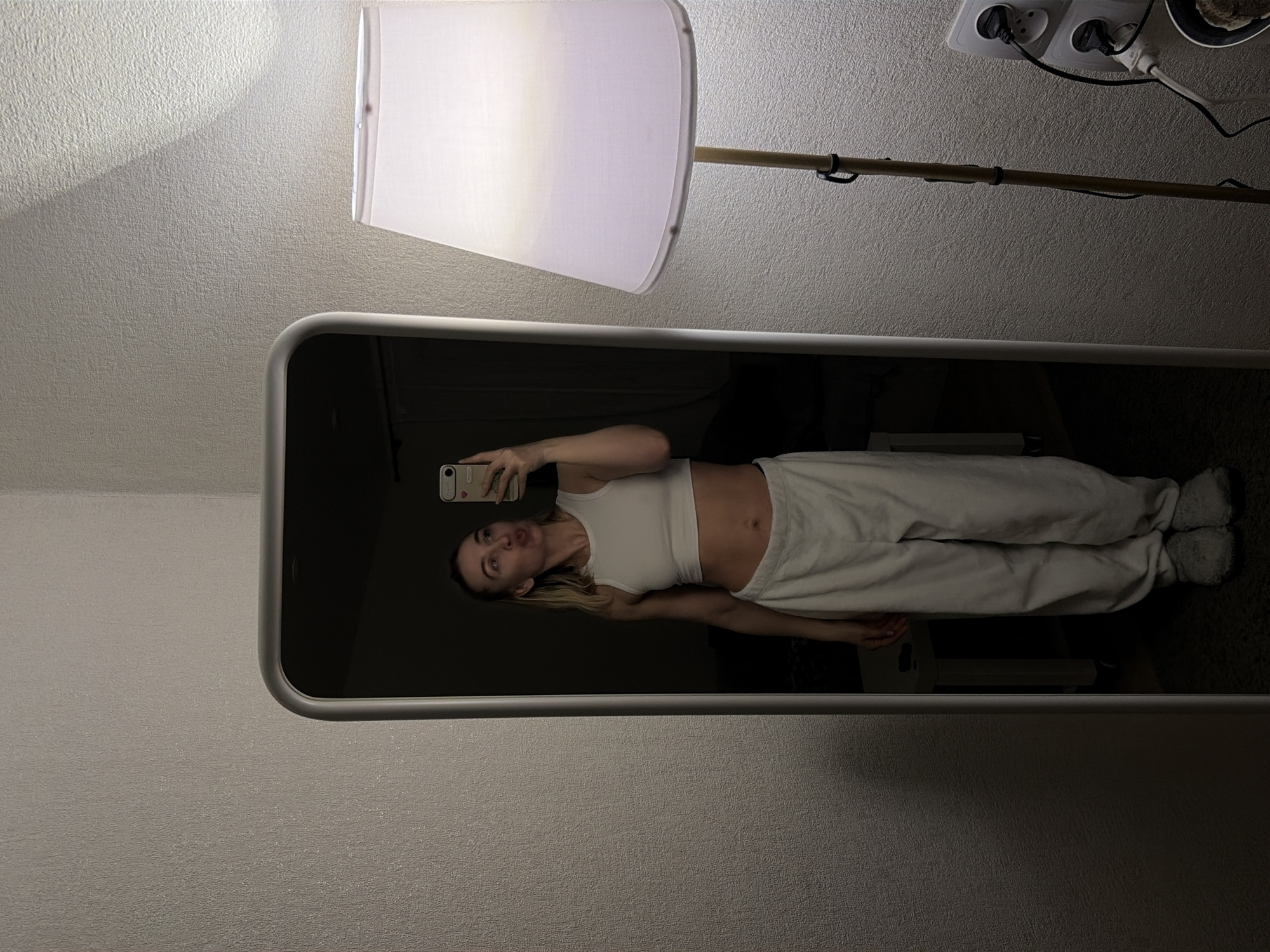 A female subject is depicted in a full-length mirror selfie taken indoors within a residential setting in Chaikovsky, Russia. The Caucasian woman, with light-colored hair, wears a white cropped tank top, light-colored loose trousers or sweatpants, and grey slippers. Her midriff is exposed. She holds a light-colored smartphone in her right hand, angled towards the mirror to capture the image, and her lips are pursed.
The mirror has a white or light-colored rounded frame and occupies the left portion of the image. To the right of the mirror, a floor lamp with a white conical fabric shade provides illumination, casting light onto a textured, light-colored wall. Below the lamp, a stacked double electrical outlet (European type) is visible, with two black power cords plugged in. The mirror's reflection behind the woman shows a darker room, partially revealing a small, light-colored table and a dark carpet or rug on the floor.