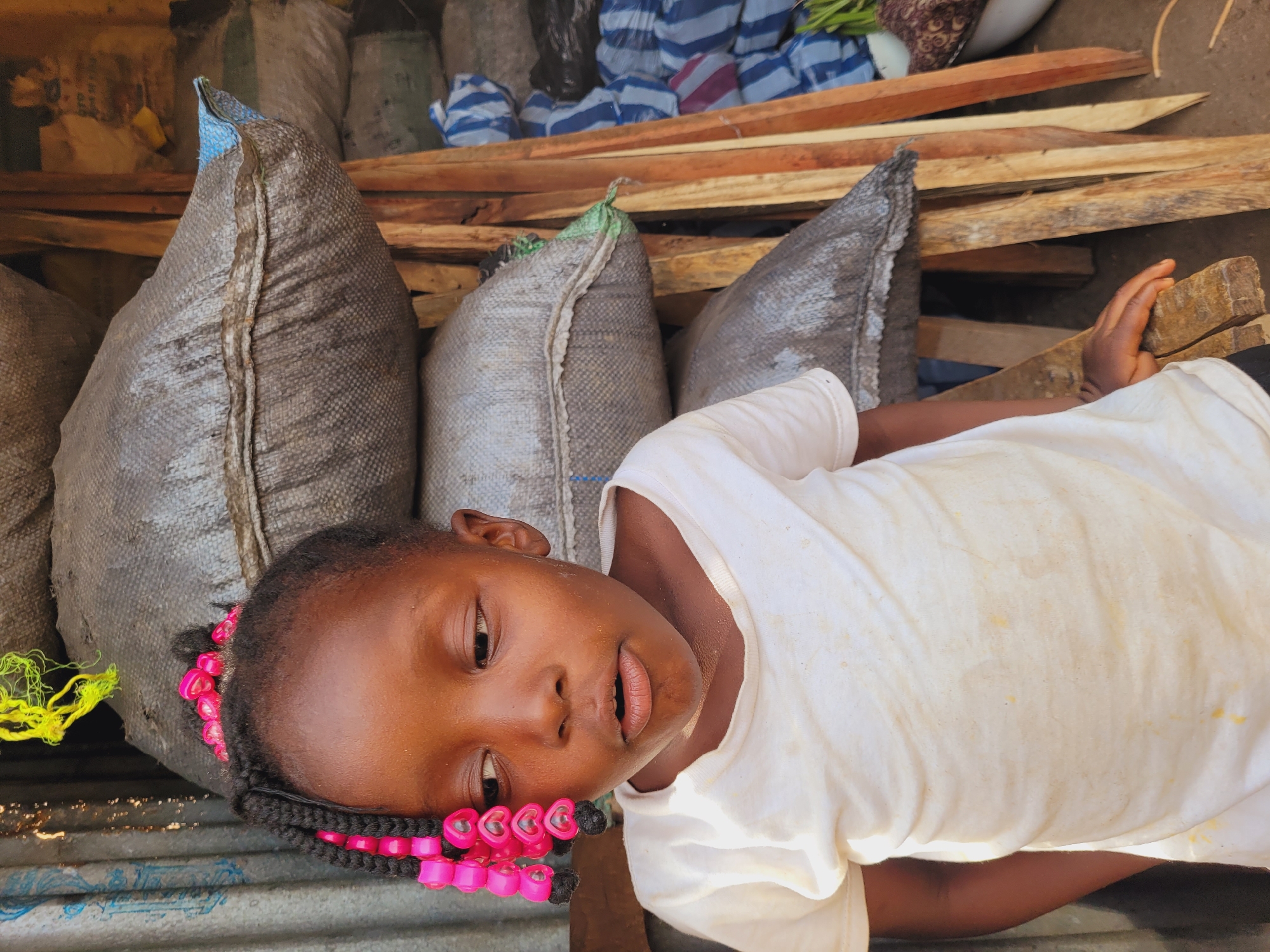 A young, dark-skinned child lies on corrugated metal next to several large, gray sacks in Bukuru, Nigeria. The child, who is wearing a white t-shirt and has braids adorned with pink heart-shaped beads, looks up towards the camera. Wooden planks and assorted bags are stacked in the background. The scene is outdoors and appears to be in a storage or market area.