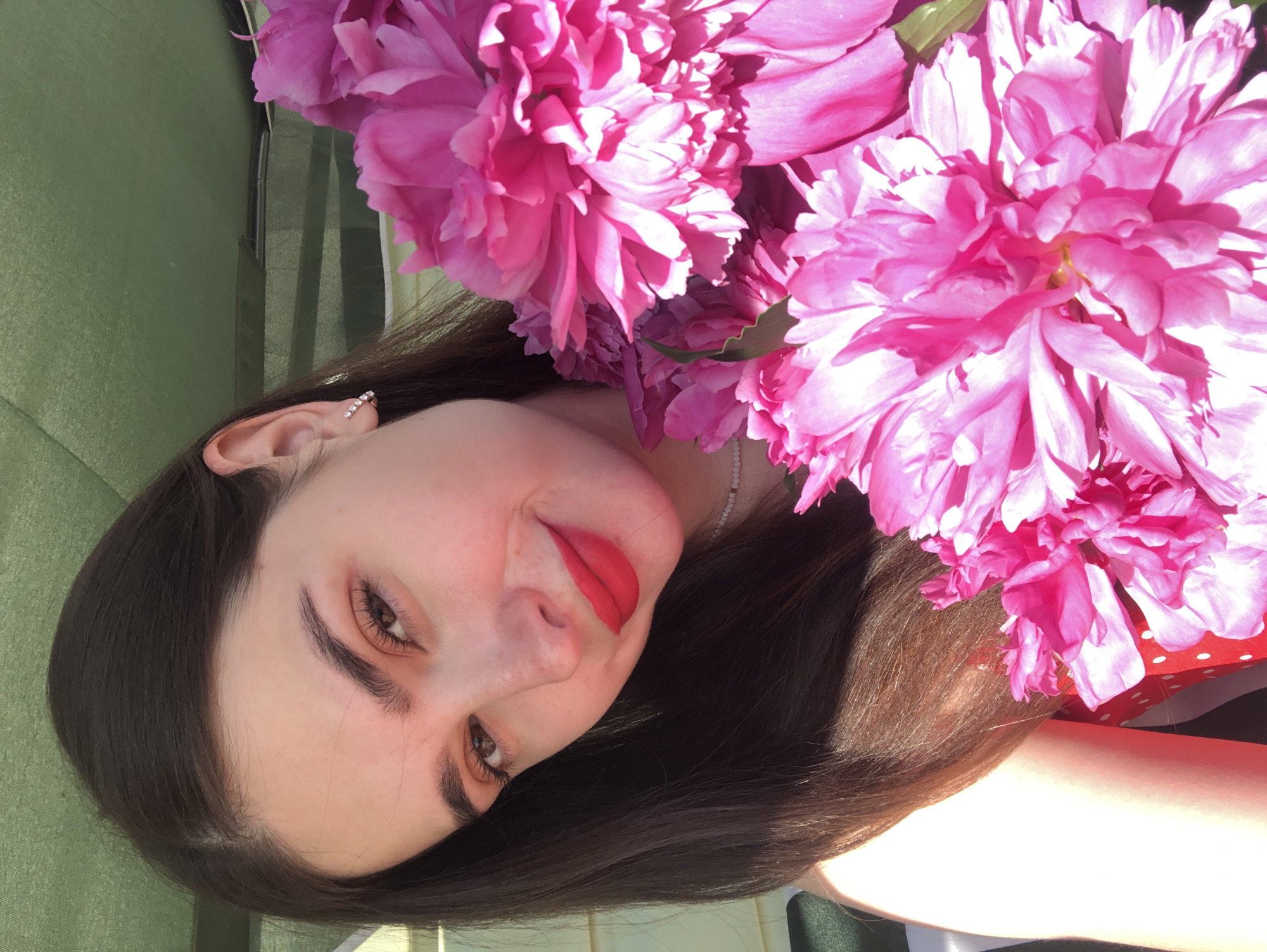 A young woman with long, dark hair and fair skin is photographed from the chest up, holding a bouquet of large, vibrant pink peonies. She is wearing bright red lipstick and a subtle pearl necklace. Her head is tilted slightly to her left, and she is looking directly at the camera with a gentle smile. The background appears to be an outdoor setting, possibly a garden or patio, with a green canvas or tarp visible overhead. The lighting suggests it is daytime, with sunlight casting shadows and highlighting the woman's features and the flowers. No specific location within Noviy, Russia can be determined from the visual information provided.