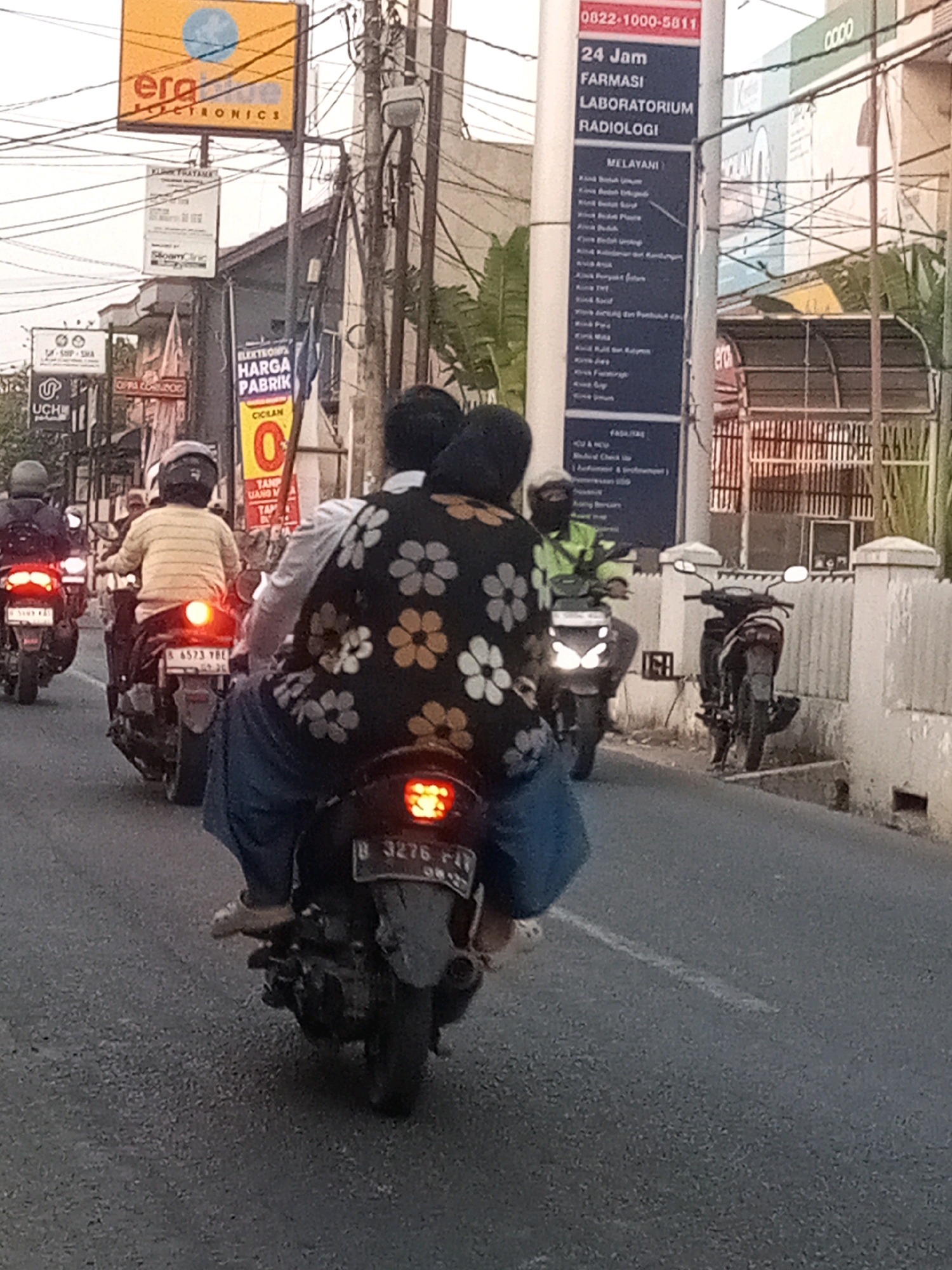 The image captures a street scene in Bekasi, Indonesia, likely during the daytime, given the overcast sky. In the foreground, two individuals are riding on a motorcycle, with the passenger in the back wearing a black shirt adorned with a floral pattern. The driver is wearing a dark head covering. The motorcycle's license plate is partially visible, reading "B 3276 F1Y".
Several other motorcycles are present on the road, indicating traffic. One motorcycle in the mid-ground has a license plate that reads "B 6573 YBE". The environment features buildings with signage, including one that says "ERA ELECTRONICS" and another for "24 Jam FARMASI LABORATORIUM RADIOLOGI". Overhead, utility poles and wires are visible, typical of urban infrastructure. The road surface appears to be asphalt. The overall atmosphere suggests a busy urban street.