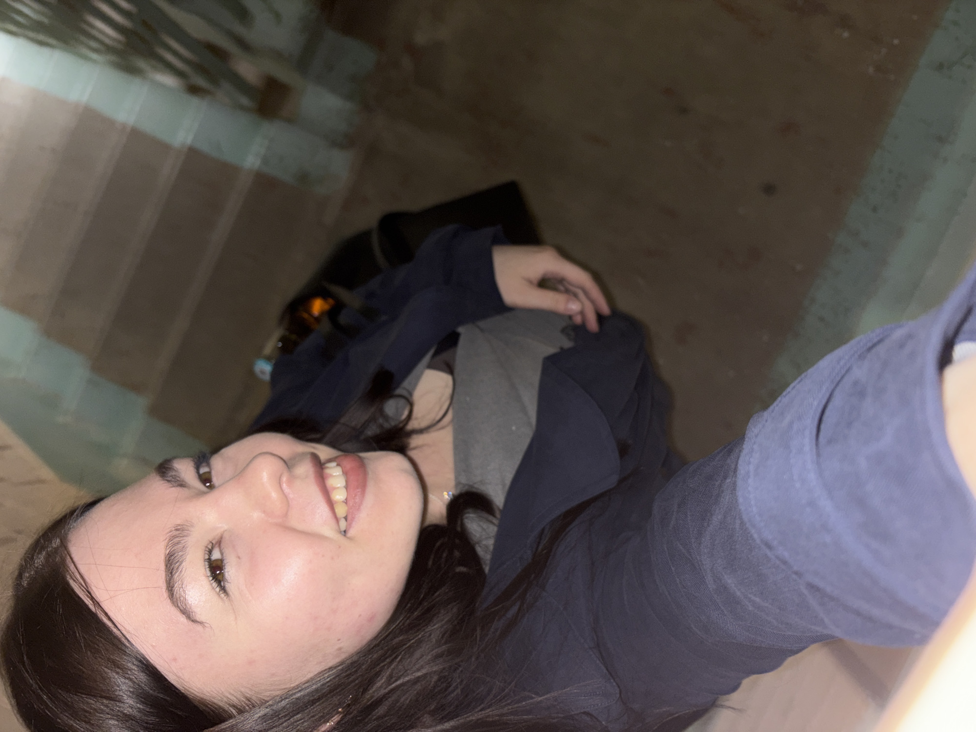 A young woman with long, dark hair, wearing a dark blue jacket and a gray shirt, is seated. She is indoors. A black bag sits behind her. The wall has stripes of light green and brown. The location is potentially Perm, Russia. No specific landmarks are identifiable.