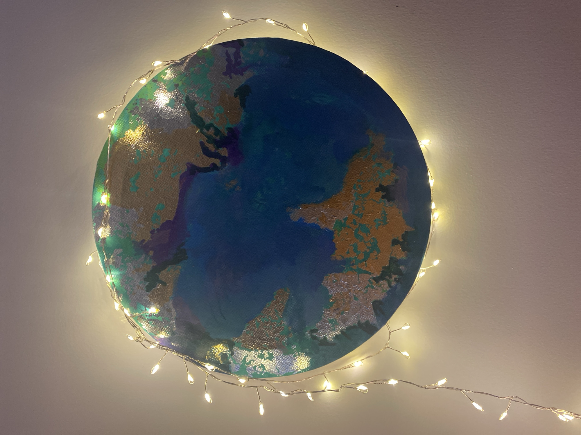 A circular, painted depiction of the earth is displayed on a light-colored wall, surrounded by a string of small, warm-toned fairy lights. The painting is a mixed-media artwork featuring continents rendered in green, gold, and silver tones, with oceans in shades of blue and purple. The light string encircles the painting, cascading down the wall slightly on the bottom right. The location cannot be determined from the image.