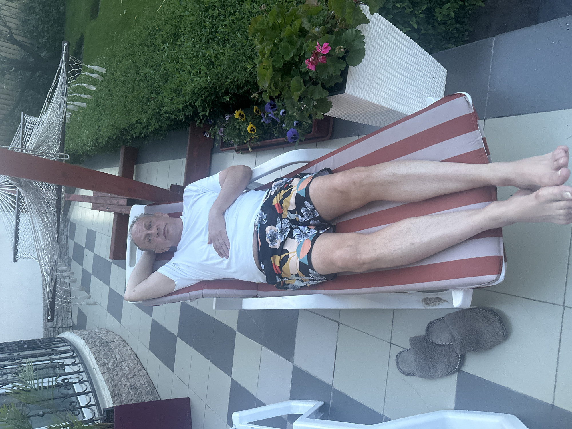 The image shows a man lying on a red and white striped lounge chair on a checkered patio. He is wearing a white t-shirt and colorful patterned shorts. His left arm is behind his head, and his right hand is resting on his stomach. To the left of the chair are a pair of grey slippers. In the background, a hammock is visible, strung between two wooden posts, and a hedge of green bushes borders the patio. Flower pots with various flowers are placed near the hedge. The patio appears to be part of a residential area, possibly a backyard or a terrace.