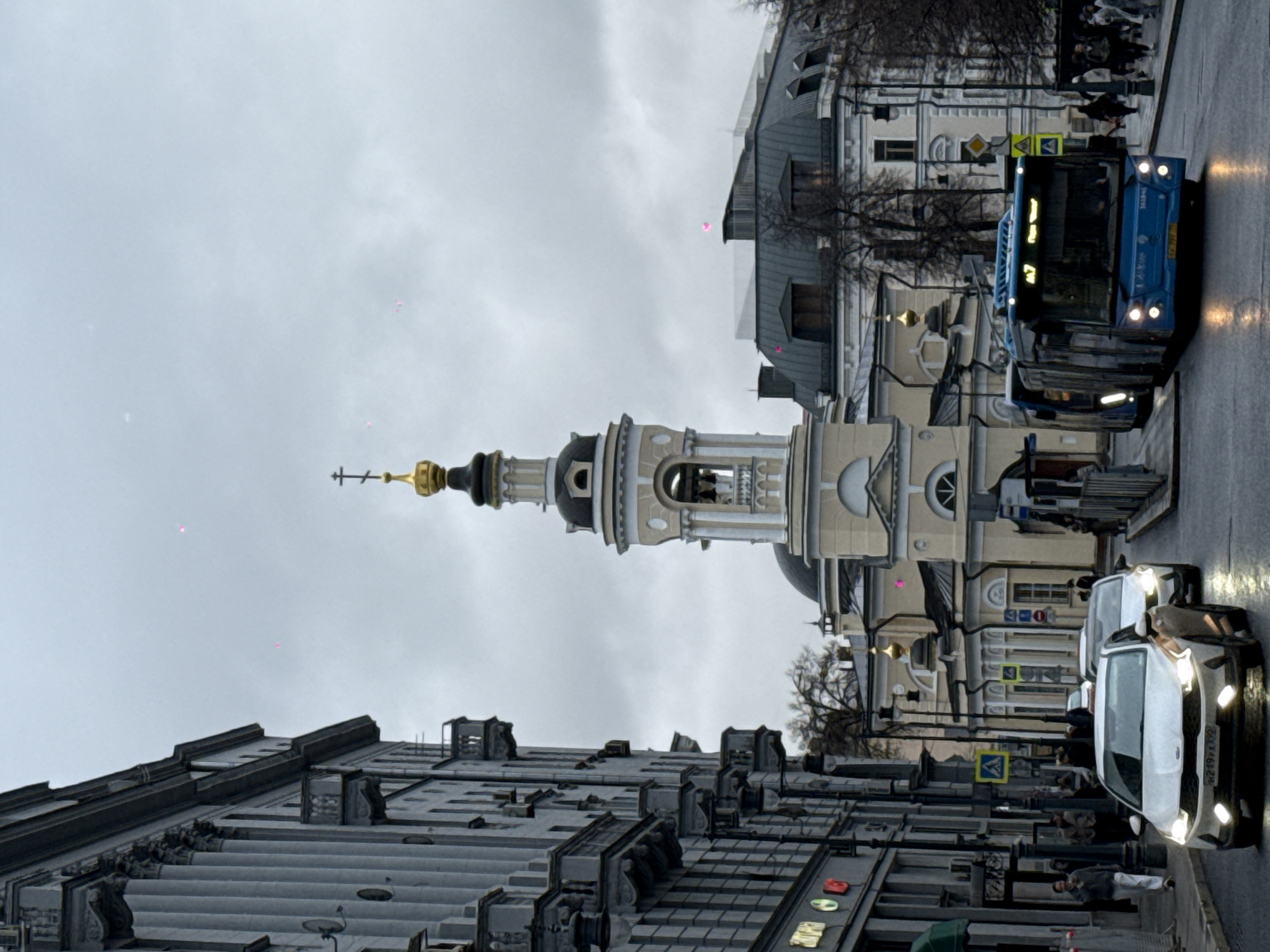 The image captures a street scene in Moscow, Russia, featuring a tall, ornate church steeple. The steeple, with its golden dome and cross, dominates the skyline. Buildings with intricate facades line the street, showing architectural details characteristic of Moscow. Vehicles, including a bus and cars, move along the wet street. Pedestrians are visible on the sidewalks. The sky is overcast, suggesting a gloomy day.