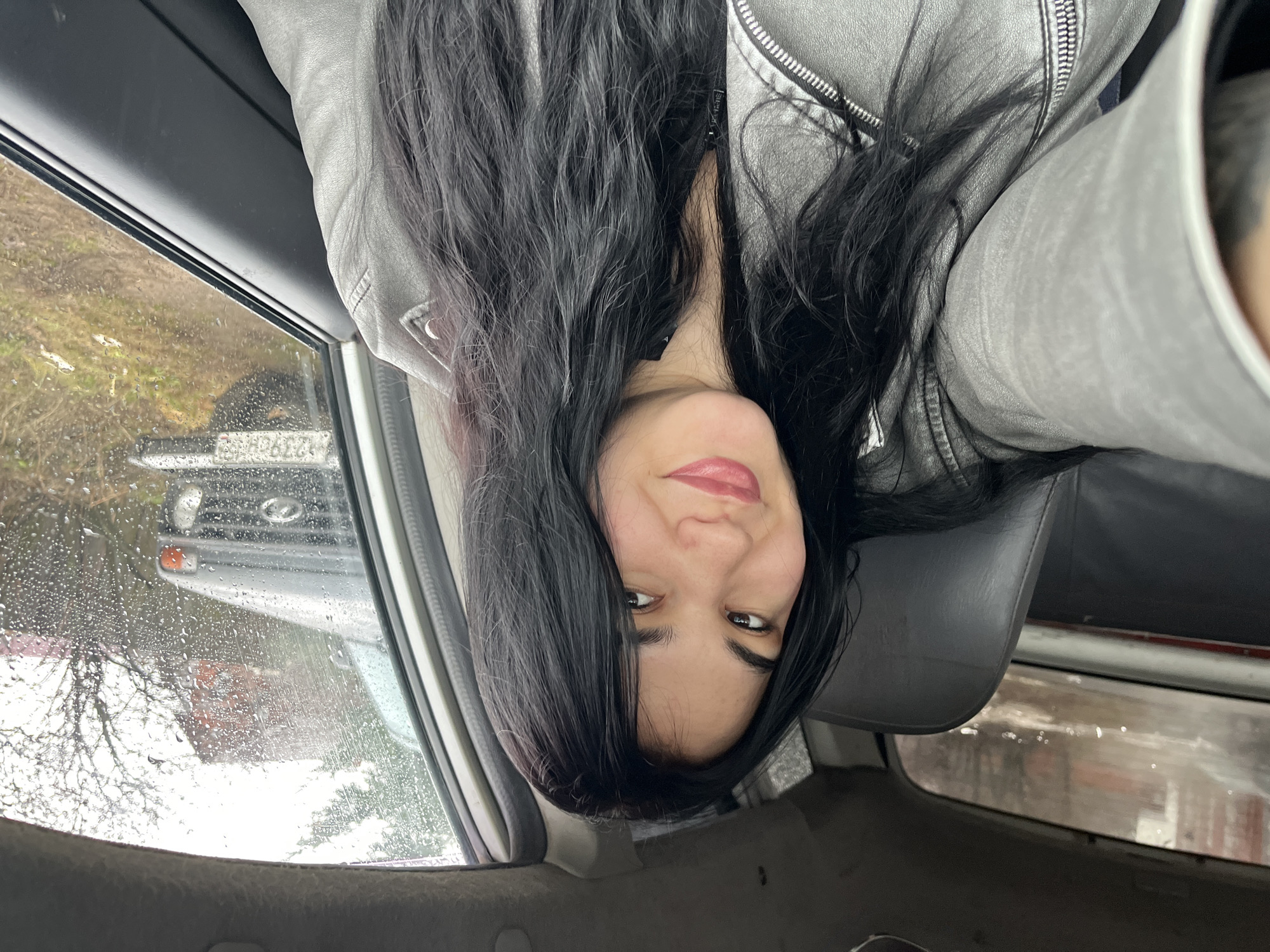 The image is a selfie taken inside a vehicle, likely a car, during rainy weather. The subject is a woman with long, dark hair, wearing a gray jacket. The interior of the car is visible, including the seats and window. Through the rain-streaked window, a blurred view of the outside world is visible. In the background, another vehicle, possibly a Lada, can be seen. The license plate number of the car is partially visible.
Based on the license plate number, the location is Chaikovsky, Russia. The scenery outside the window suggests a typical urban landscape in the region, characterized by buildings, trees, and roads. The rain creates a wet and slightly gloomy atmosphere.