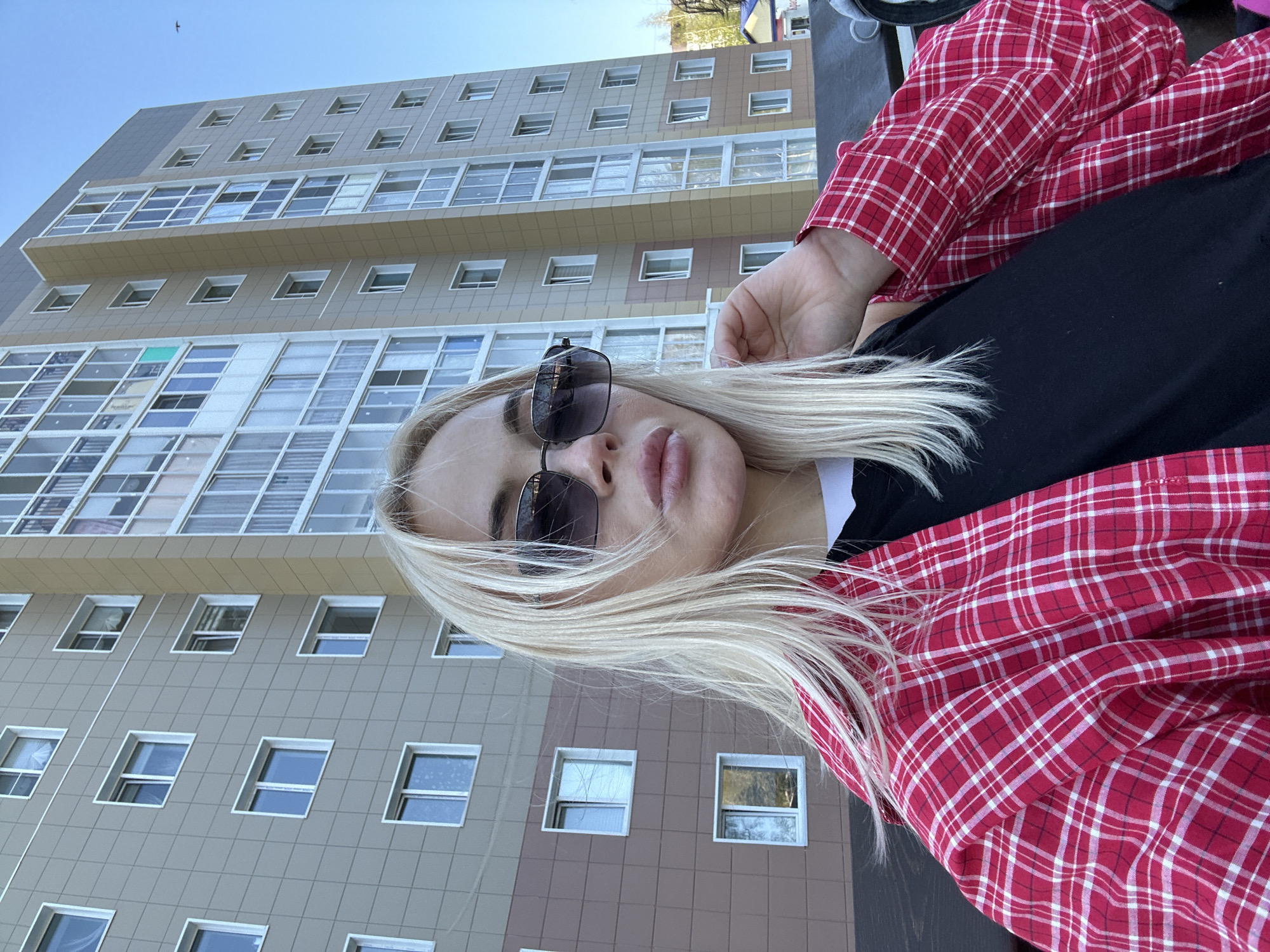 A woman with blonde hair and sunglasses is outdoors, wearing a red and white plaid shirt over a black top. Behind her is a multi-story building with numerous windows. The building is located in Megion, Russia.