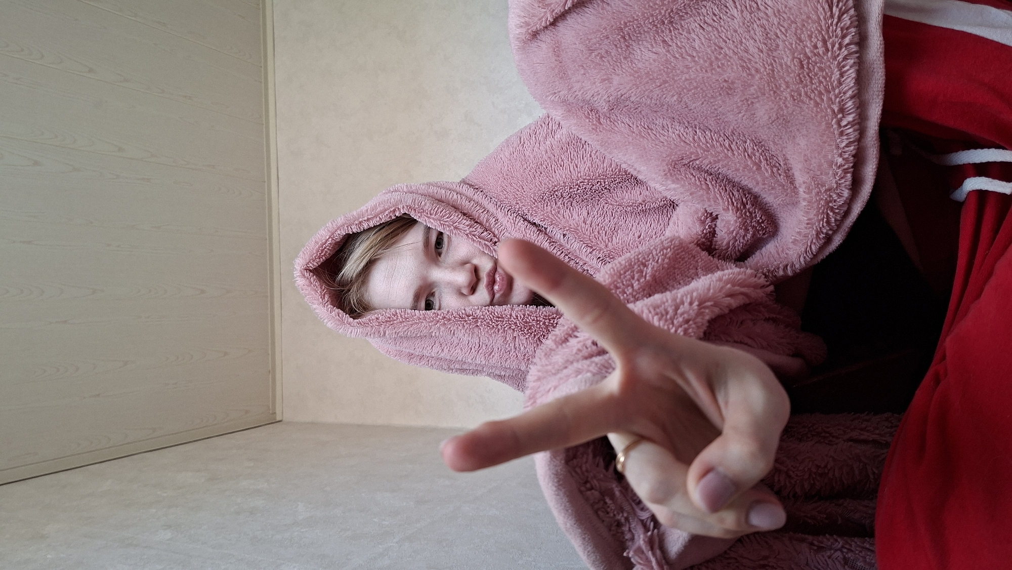 A young person, possibly female, with fair skin and light blonde hair, is facing the camera. They are wearing a fluffy, hooded, pink bathrobe or similar garment. They are holding their hand up, displaying a "peace" or "victory" sign with their index and middle fingers. The background is a sparsely furnished room with a light-colored wall and what appears to be a light beige or grey floor. A white door or panel is visible on the left side of the frame. Part of a red object, possibly a garment, is seen at the top right corner of the image. The photo appears to be taken indoors with natural lighting. There is no specific information to confirm the location as the city of Большой Букор, Russia.