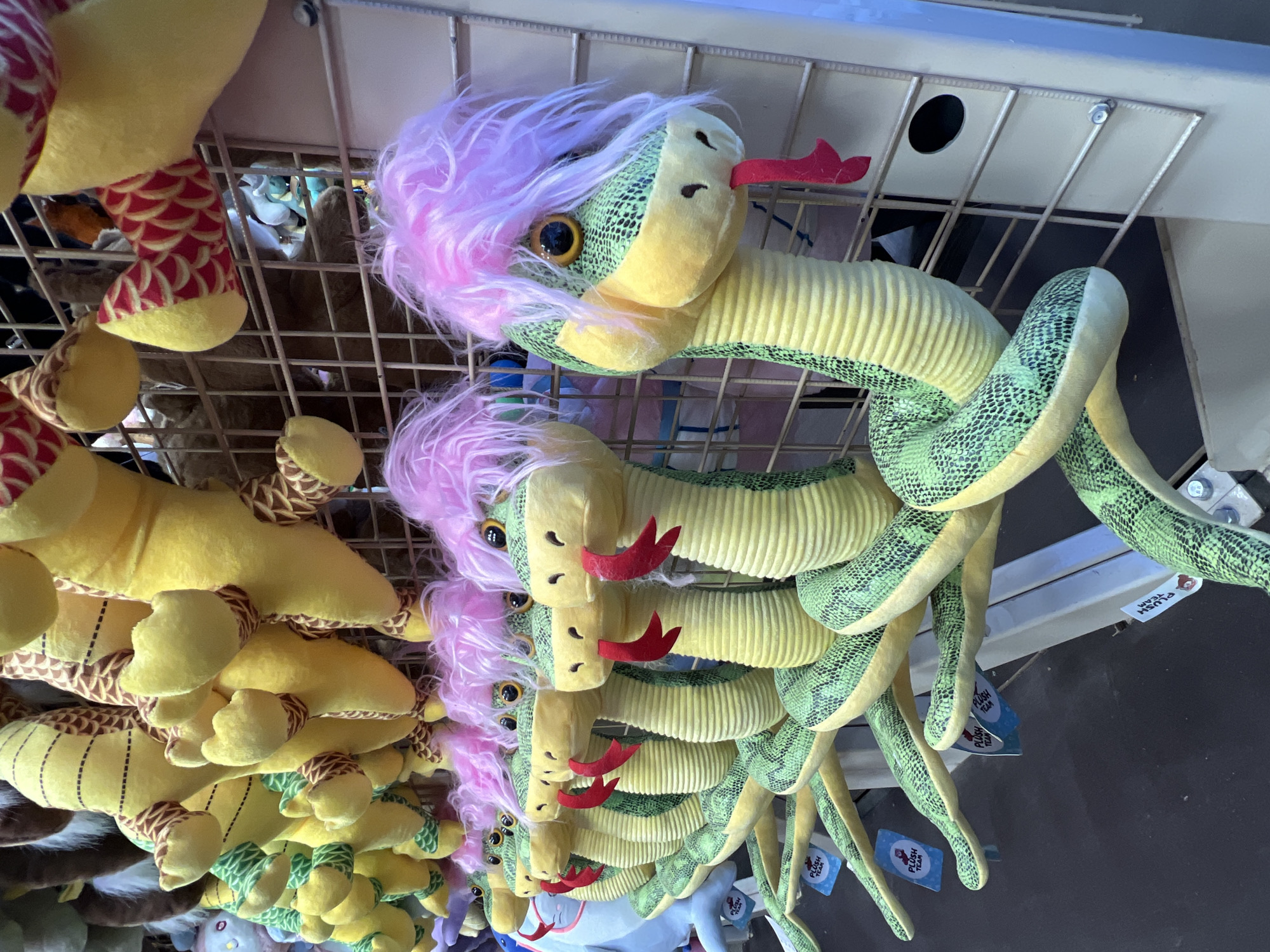 A slightly rotated, eye-level shot depicts a merchandise display of plush toy snakes, likely at a retail location. The snakes are yellow and green, with pink hair, and red forked tongues. They are hanging from a wire rack. The snakes are suspended by their bodies, with their tails dangling down. Some of the snakes have tags attached. The presence of Russian text or landmarks is not visible, so locating the image in Cherepovets, Russia, is impossible.
