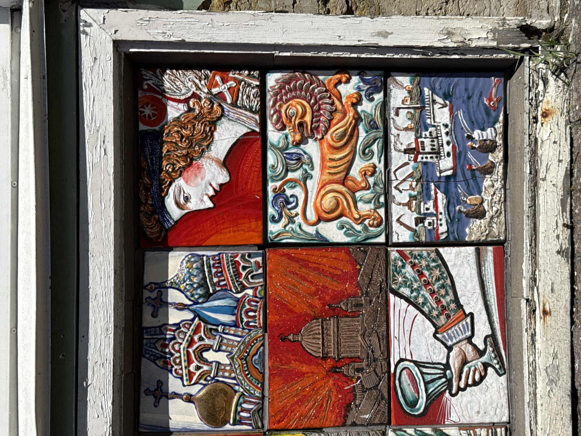 The image displays a grid of six ceramic tiles set into a weathered, peeling white wooden frame. The tiles are arranged in two columns and three rows, forming a decorative mosaic. The top left tile features a colorful depiction of Russian Orthodox churches with distinctive onion domes and crosses. The top right tile shows a profile of a stylized human face with curly orange hair, a blue patterned cap, and prominent red cheeks, set against a red background with some white and green embellishments. The middle left tile depicts a large dome building, likely a cathedral, with radiating orange lines behind it, against a dark, textured background. The middle right tile showcases a vibrant orange lion with a flowing mane, in profile, walking amidst green foliage. The bottom left tile presents a close-up of a hand, rendered in muted tones, holding a chalice-like goblet. The hand wears a jacket with a patterned sleeve featuring small stars and red buttons. The bottom right tile depicts a maritime scene with several ships on a blue sea, smoke emanating from their funnels. Small figures are shown on a rocky shoreline, and a red fish swims in the water. The tiles are installed within a rough, aged wooden frame that shows significant wear, paint chipping, and some rust stains on the lower part. The background outside the frame suggests an outdoor setting, with the texture of a stone wall visible on the right. The overall impression is of an outdoor art installation, possibly a mosaic window or a facade decoration. Given the architectural style of the churches in the top left tile, the mosaic is likely located in Saint Petersburg, Russia, a city renowned for its numerous churches and historical buildings.