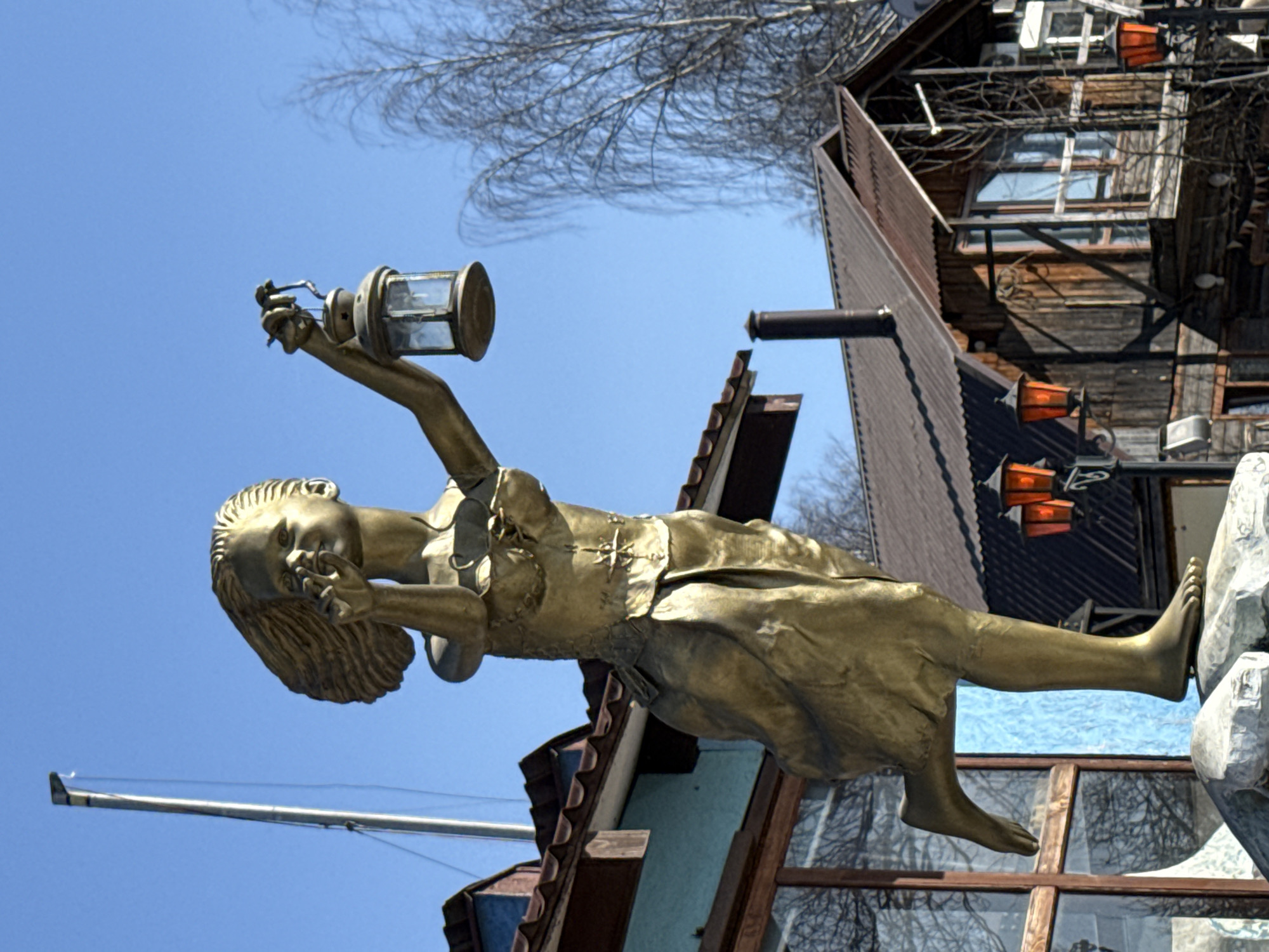 A statue of a girl holding a lantern is featured in this full, rotated shot. The statue is bronze and shows a girl in simple clothing with wavy hair, raising a lantern with one hand. The statue is positioned on a building, with architectural details such as the roof and wooden window frames visible in the background. The background shows the sky and bare tree branches. This statue is part of the "Children of a Different World" sculpture composition installed in the Izmailovo Kremlin cultural complex, which is located in Moscow, Russia.