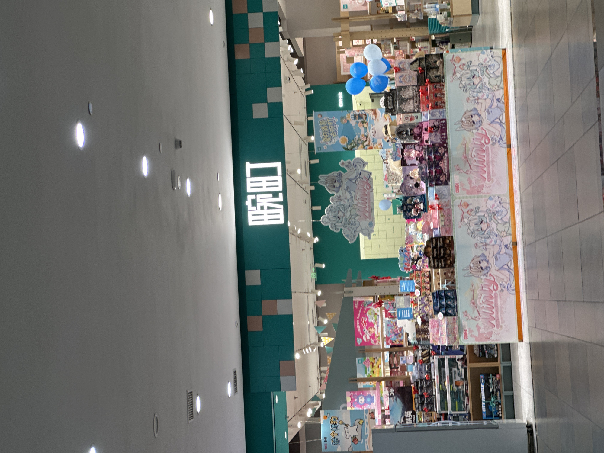 The image shows the entrance to a store with a green and teal facade. A sign with white Chinese characters, "晓町" (Xiǎo Dīng), is prominently displayed on the green section of the facade. Below the sign, the store interior is visible, filled with shelves stocked with various merchandise, including what appear to be toys and collectibles. Several colorful banners and displays are set up in front of the shelves, featuring anime-style characters and graphics. Some blue and white balloons are tied to a display near the right side of the entrance. The floor in front of the store is tiled with a grey and white pattern. The lighting suggests an indoor environment, likely a shopping mall or commercial area. The scene is set in the city of Chongqing High-tech Zone, China.