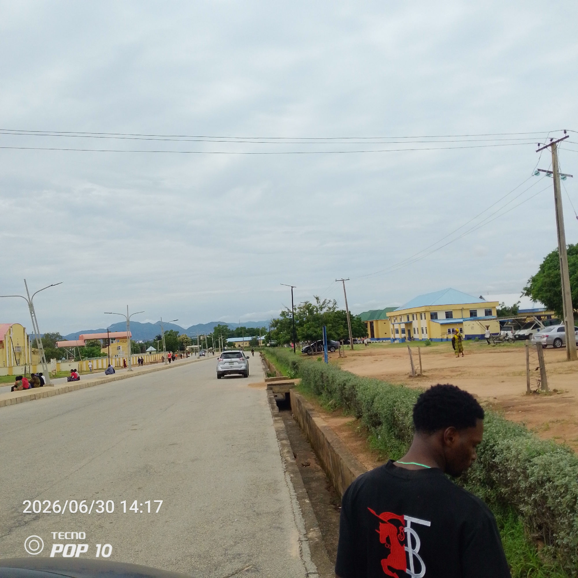 This media file depicts a street scene in Jalingo, Nigeria, on a cloudy day. The foreground features a man wearing a black t-shirt with a red and white graphic design. He is looking down and away from the camera. The middle ground shows a paved road with several vehicles, including a silver car driving away from the viewer and a parked white van. People are visible along the sidewalk and on the side of the road, some sitting and others walking.
The background reveals a cluster of buildings with yellow and blue facades, suggesting an institutional or commercial area. Mountains are visible in the distance, softened by the overcast sky. Lampposts line the street, and utility poles with electrical wires are present. The overall atmosphere is casual, with people engaged in daily activities.
Visible text includes a timestamp in the lower left corner: "2026/06/30 14:17" and a watermark that reads "TECNO POP 10". The weather appears to be cloudy, with diffused light. The scene conveys a sense of ordinary life in a Nigerian urban environment.