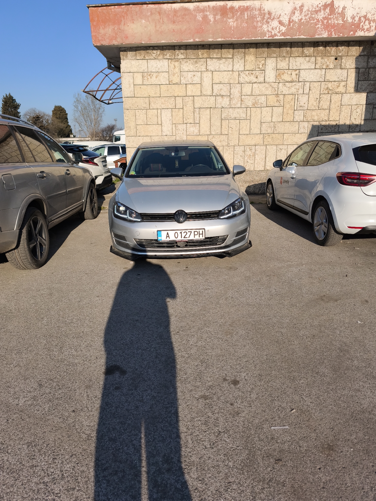 The image shows a silver Volkswagen Golf parked between a silver Volvo SUV and a white Renault Clio electric vehicle in an outdoor parking lot. The Golf has a license plate that reads "A 0127 PH". The parking lot is asphalt and appears to be located near a building with a stone block facade. The shadow of a person, presumably the photographer, is visible in the foreground. The background shows other parked vehicles and some sparse trees. Based on the license plate format and other environmental clues, the location is likely in Burgas, Bulgaria.