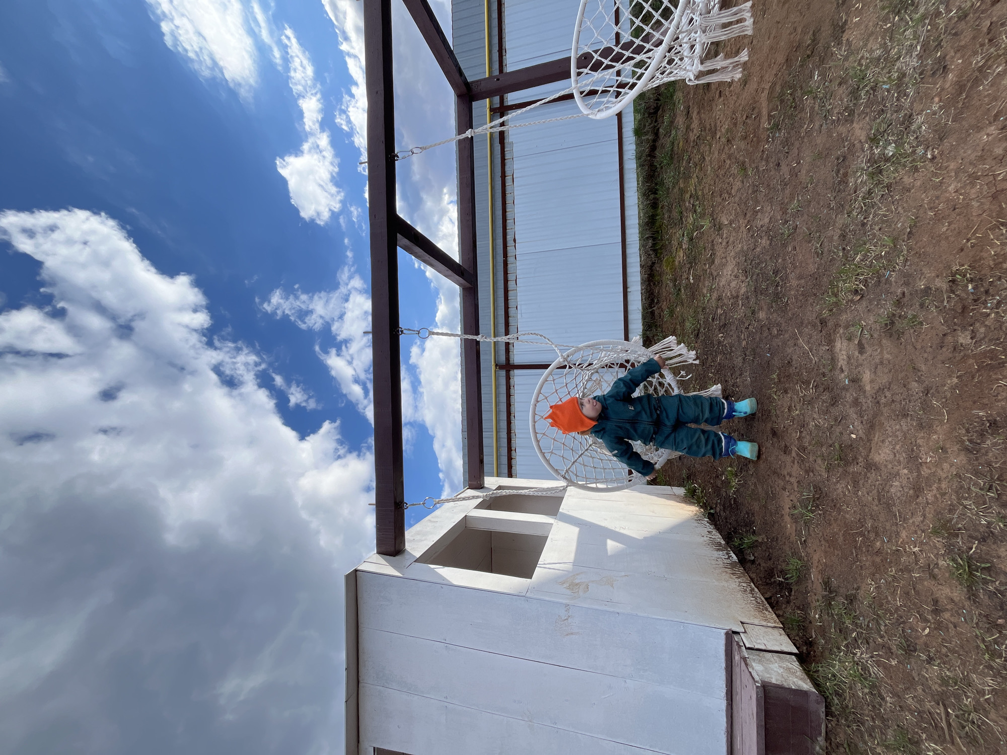 A child wearing an orange hat, a dark green jumpsuit, and light blue boots is sitting in a white macrame hanging chair in the city of Chaikovsky, Russia. The chair hangs from a metal beam above a white building with a window. The sky is visible above, with blue patches and white clouds. The ground next to the building is brown and sparsely covered with vegetation.