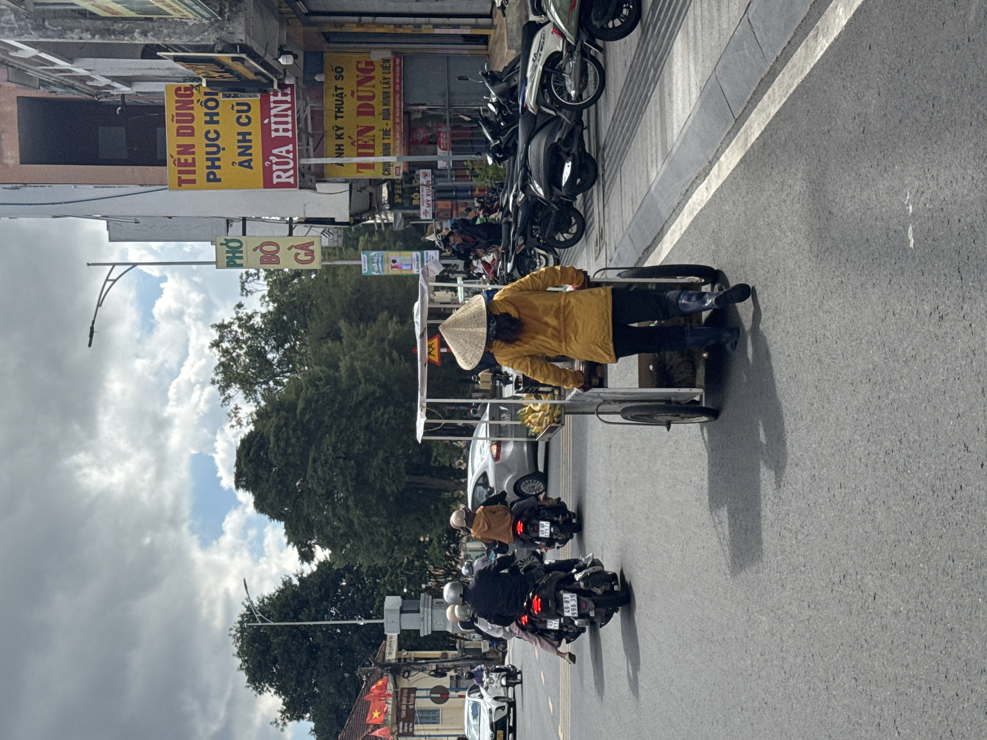The image depicts a street scene in Phường Xuân Hương - Đà Lạt, Vietnam, on a cloudy day. In the foreground, a woman wearing a conical hat and a yellow jacket pushes a cart with bananas. She is walking away from the camera on the right side of the road. Several motorbikes are visible on the road, with riders wearing helmets and protective clothing. On the left side of the road, a white car is parked, and another car is driving in the distance. Buildings with signs in Vietnamese line the right side of the street. The overall environment suggests a bustling urban setting with common modes of transportation.