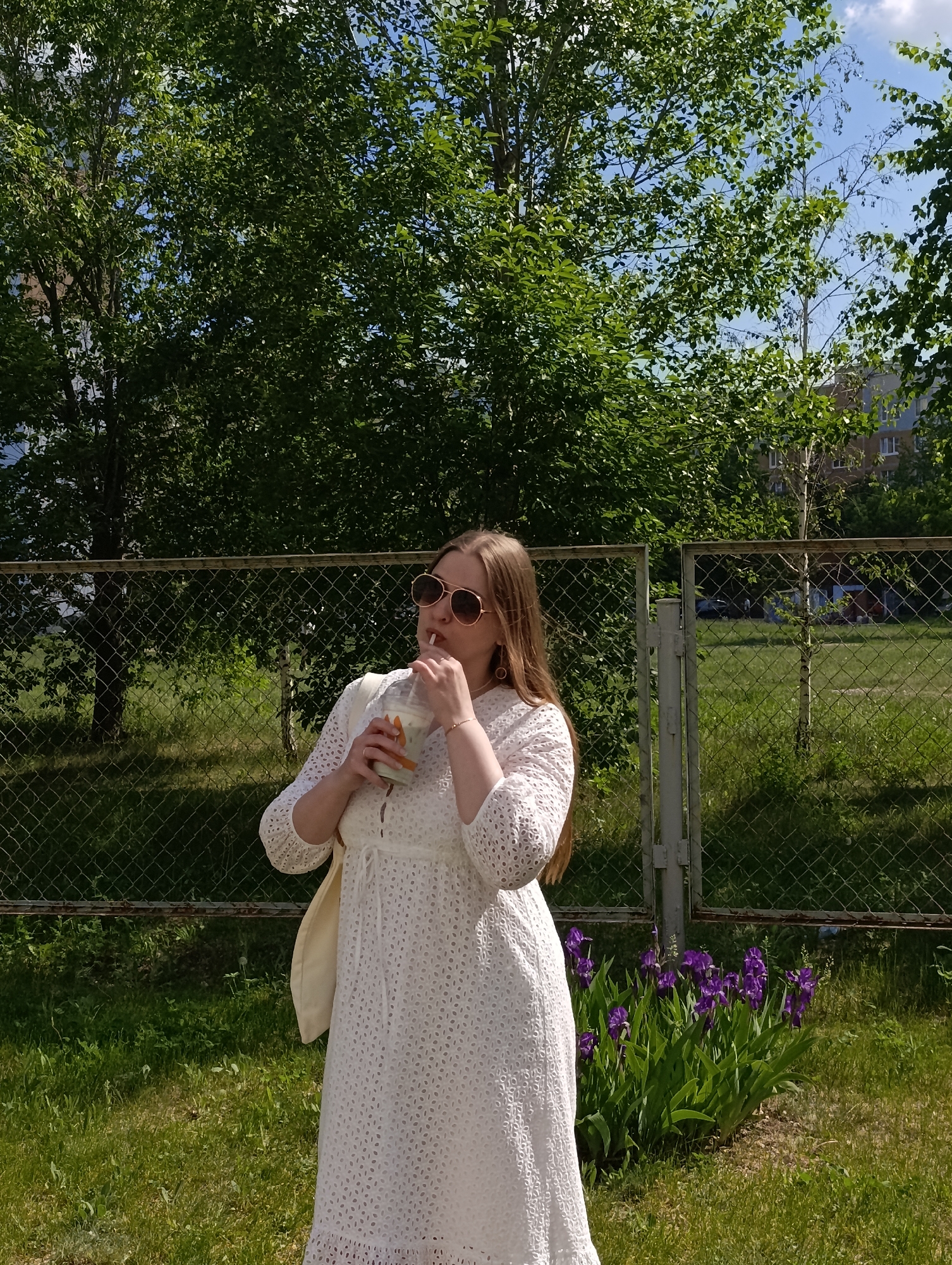 The image shows a woman standing outdoors in a grassy area, likely a yard or park in the city of Unknown, Unknown. She is wearing a white, eyelet-patterned midi dress, sunglasses, and has long brown hair. She is holding and drinking from a clear plastic cup with a straw. A light-colored bag is slung over her shoulder. Behind her is a chain-link fence, greenery, and a patch of purple irises. In the background, trees and a building are visible. The weather appears sunny.
