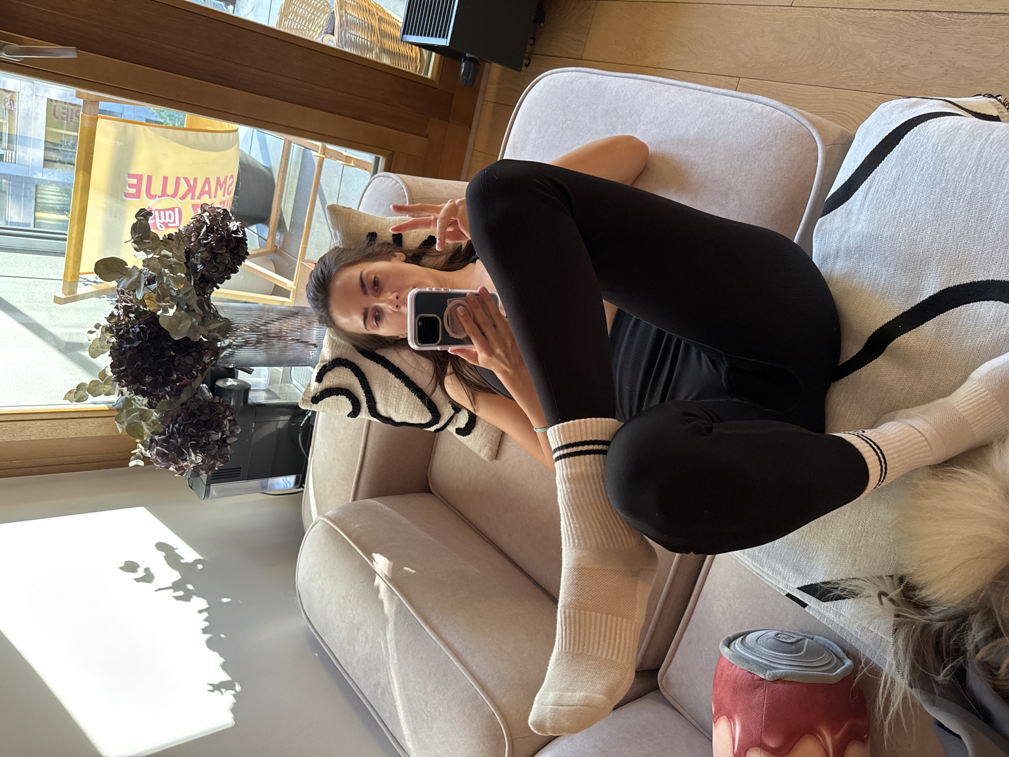A woman wearing black leggings and white socks with black stripes is lying on a light-colored couch. She is holding a cell phone in her left hand and making a peace sign with her right. The woman is positioned in an apartment in an unknown city. A vase of flowers is on a table next to the couch, and a window in the background provides natural light. A decorative pillow is also visible. A can shaped pillow in the bottom right corner of the image.