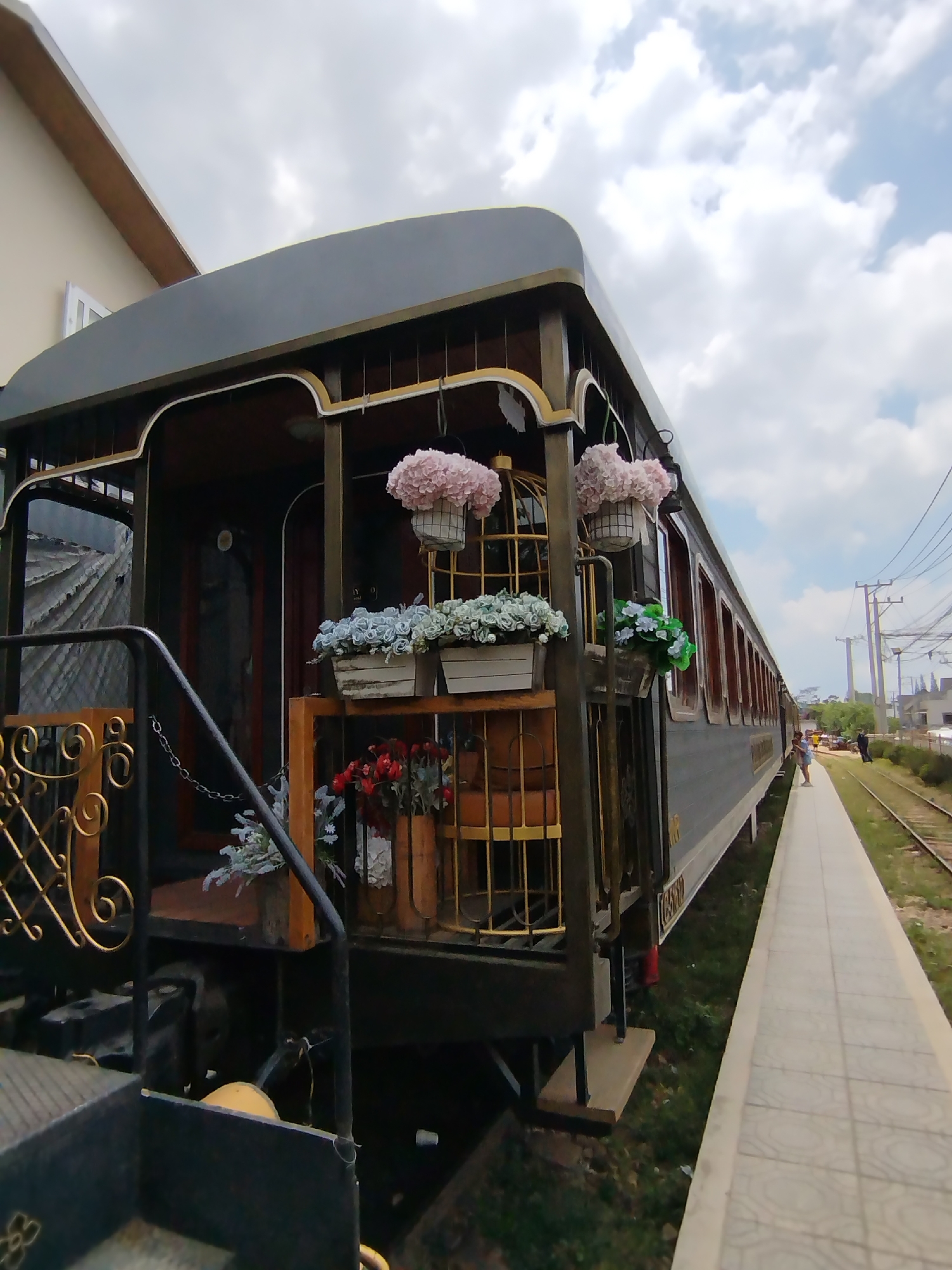 A vintage train car, adorned with flower boxes, sits on a track adjacent to a tiled platform in Lộc Quý, Vietnam. The train car is painted a dark color with gold accents and contains a staircase for passengers to enter. Two people stand on the platform towards the end of the train car. Power lines, utility poles, and buildings are visible in the background.