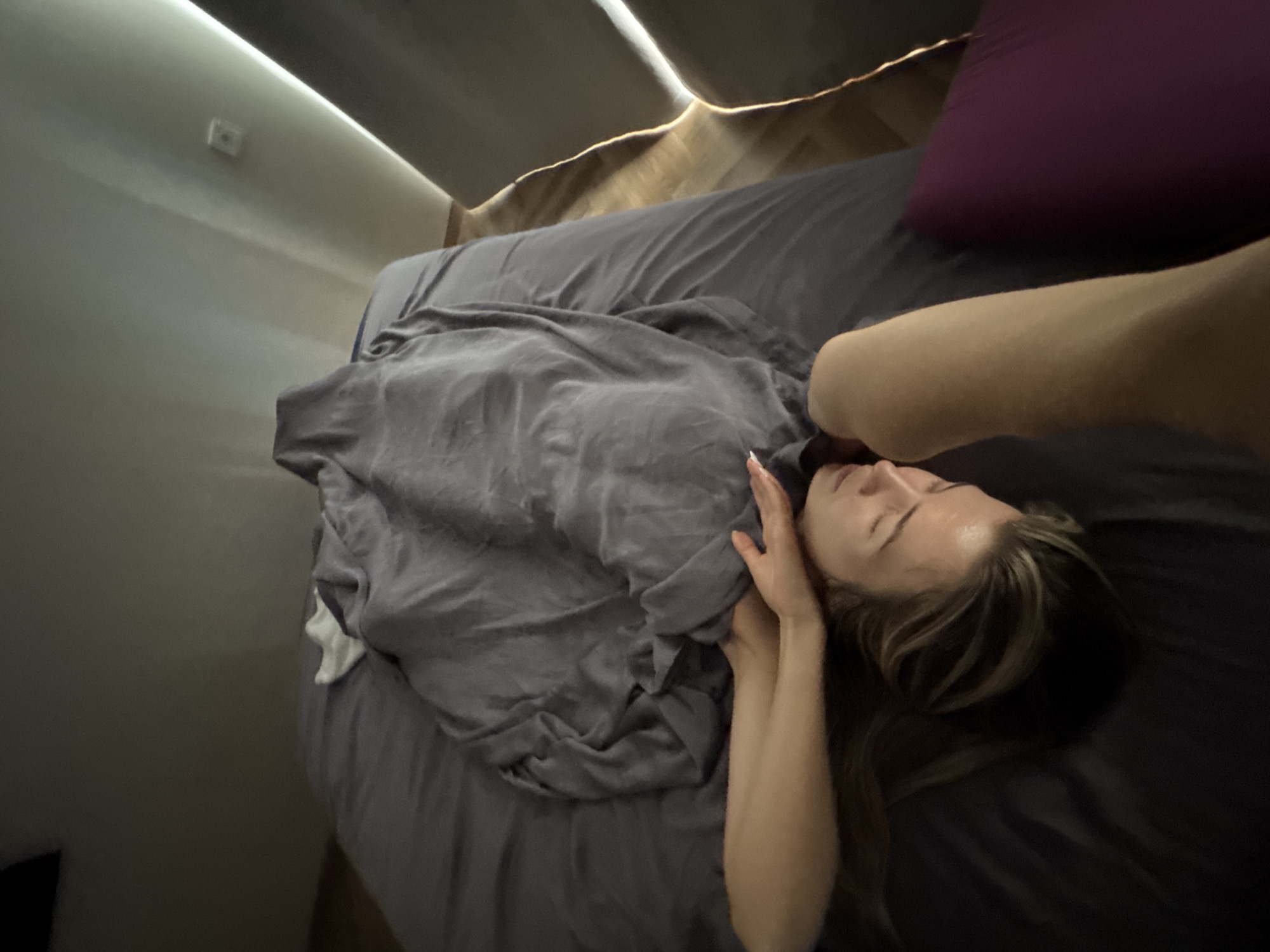 A person is lying on their side in a bed, with their face turned away from the camera and their eyes closed. They are covered by a gray blanket, which is draped over them in a disheveled manner. Their right arm is extended above their head, and their hand is resting on their forehead. Their hair is long and light brown, and it is spread out on the pillow. The bed appears to be made with gray sheets and a dark gray duvet cover. In the background, a wall is visible with a light strip running across it, creating a stark contrast. The scene is dimly lit, with a soft glow emanating from the light strip. A dark purple pillow is visible at the foot of the bed.