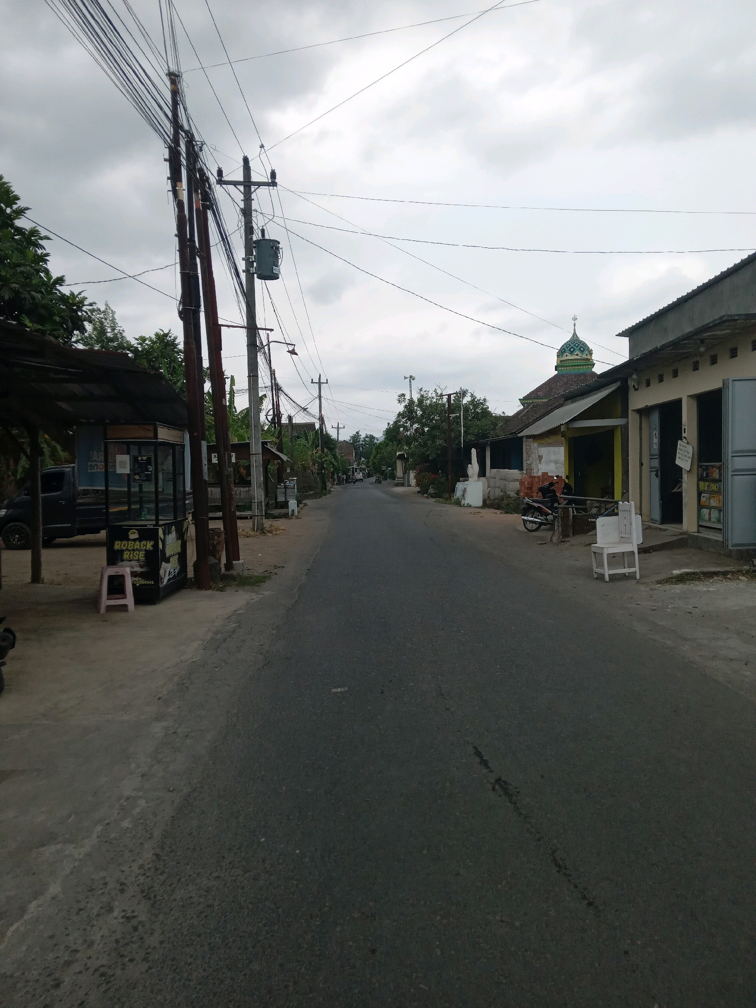 The image depicts a street scene in Banguntapan, Indonesia, under an overcast sky. The road is paved and appears to be a local thoroughfare, with a sidewalk or shoulder area on the left side. Along the left side of the street, there is a small, covered kiosk with a pink stool in front of it. The kiosk has signage that includes the words "ROBACK RISE". A dark-colored truck is parked behind the kiosk. Further down the street on the left, there are more structures and vegetation.
On the right side of the street, there are several buildings. One building has an open storefront with items displayed inside and a light-colored chair outside. Adjacent to this building, a mosque is visible with a distinctive green and white dome and a spire. A motorcycle is parked near the mosque. Various electrical poles and wires are prominent throughout the scene, indicating the presence of utility infrastructure. In the distance, the road continues, lined with trees and more buildings, suggesting a semi-urban or suburban environment. There are no people clearly visible in the image. The overall atmosphere is subdued, likely due to the cloudy weather.