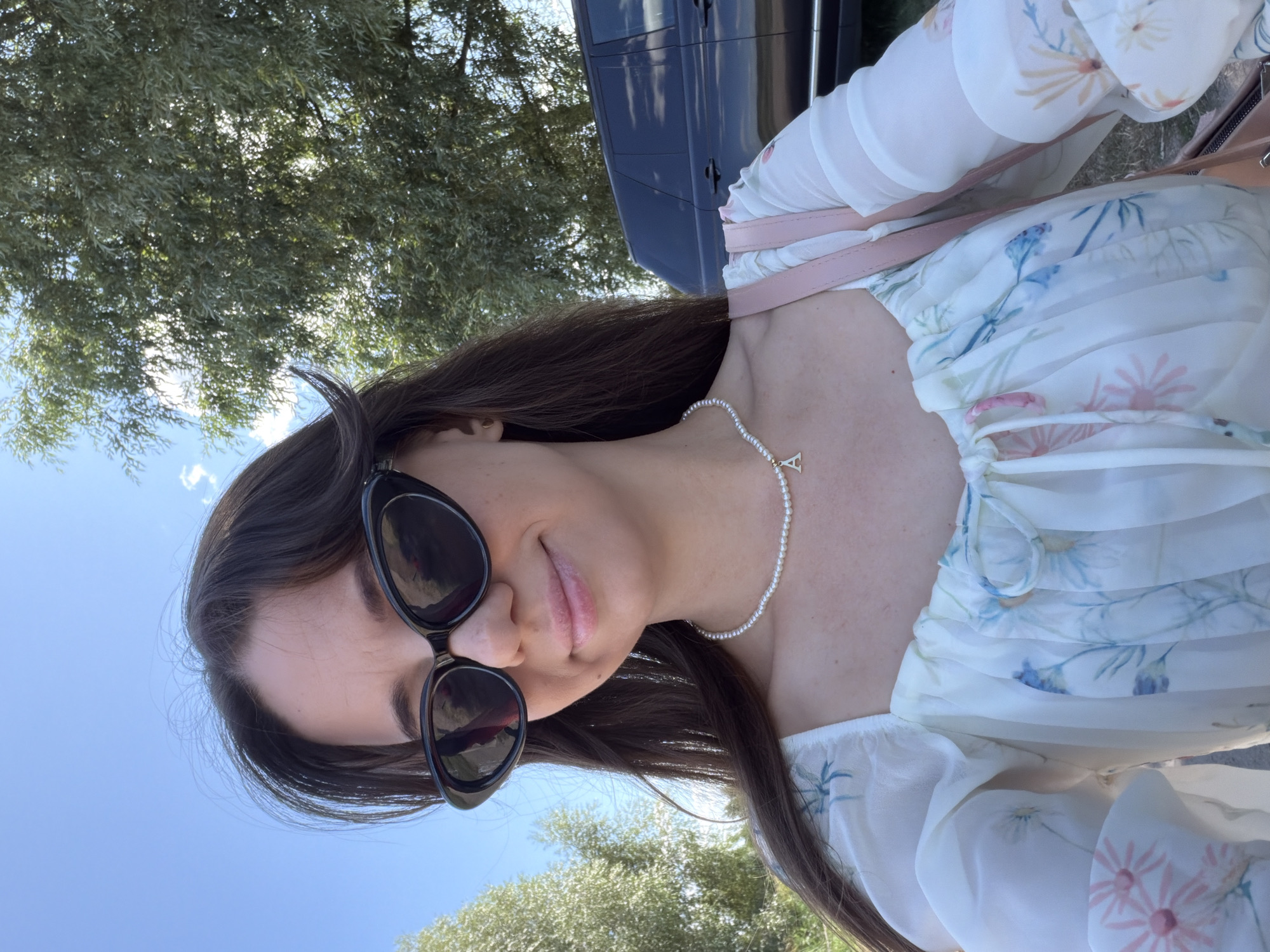 A selfie of a young woman with dark hair and sunglasses is taken outdoors on a sunny day. She is wearing a white floral-print dress and a pearl necklace with a gold initial "A" charm. Behind her, a dark SUV is parked next to a large tree. The background shows a clear blue sky with some clouds and green foliage, suggesting a rural or park-like setting outside of Severynivka, Ukraine.