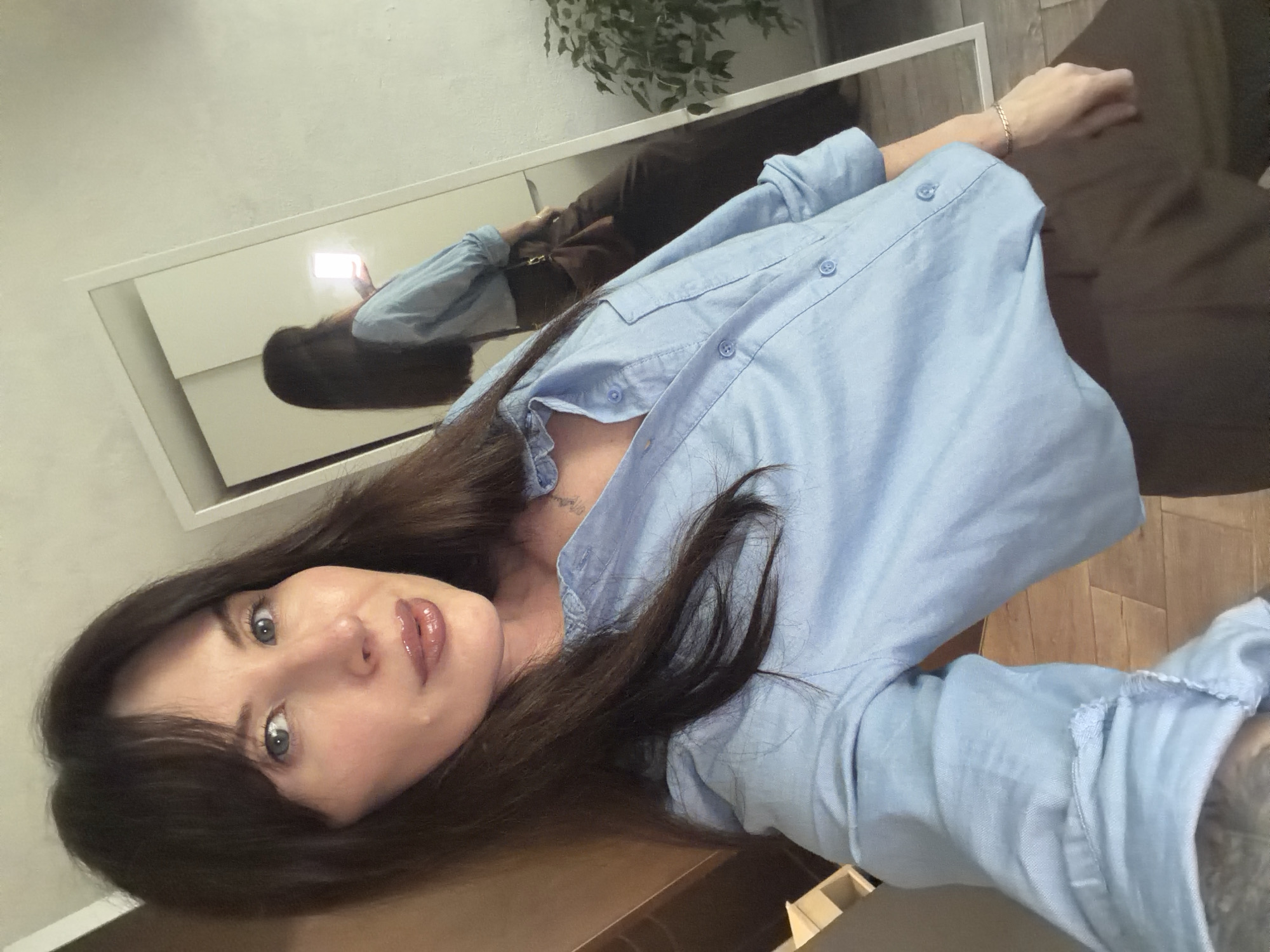 A person is taking a selfie in front of a mirror. The location appears to be indoors. The subject in the photograph is a fair-skinned person with dark hair and blue eyes. They are wearing a light blue button-down shirt. The mirror reflects their image along with a glimpse of what appears to be a brown outfit and a portion of a plant. The mirror is framed in white. The reflection shows the person holding a phone. The overall setting appears to be a room with light-colored walls. There is no indication in the image of the city Perm, Russia.