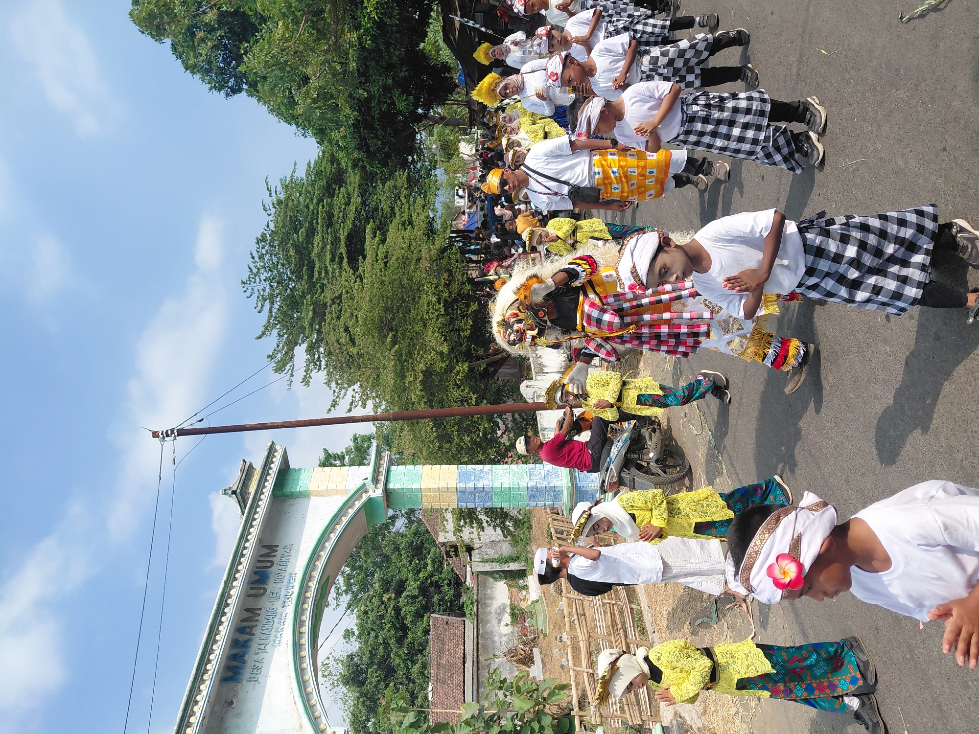 The image captures a procession on a paved road under a clear blue sky with scattered clouds. On the left, an arched gateway with the text "MAKAM UMUM DESA TALIKANDANG KEC. KOTAANYAR MAKKA THUWAK" is visible, suggesting the location is a public cemetery in Talikandang Village, Kotaanyar District, Makka Thuwak. A group of children, dressed in traditional yellow attire with headbands, are walking in the foreground, their hands clasped in prayer. To their right, a person in a red shirt sits on a motorbike, and further to the right, more children in similar attire are present. In the middle of the procession, a performer in a colorful costume, possibly a lion dance or traditional Balinese Barong, is walking. Adults in white shirts and patterned sarongs, along with others in traditional clothing, follow. The environment includes trees lining the road and buildings in the background, indicating a rural or semi-urban setting. The overall scene depicts a cultural event or parade.