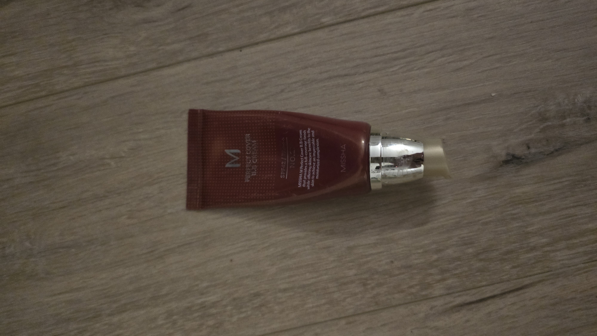 A dark red tube of MISSHA M Perfect Cover B.B. Cream, with a silver cap, rests on a wooden floor. The tube has white lettering that reads "M", "PERFECT COVER B.B. CREAM", "SPF42/PA+++", "NO.2", and a description of the product's benefits. The brand name "MISSHA" is also visible in white lettering near the bottom of the tube. The image is taken from a slightly elevated angle, focusing on the product. The environment is a plain, indoor setting with no discernible features of the city of Batumi, Georgia.