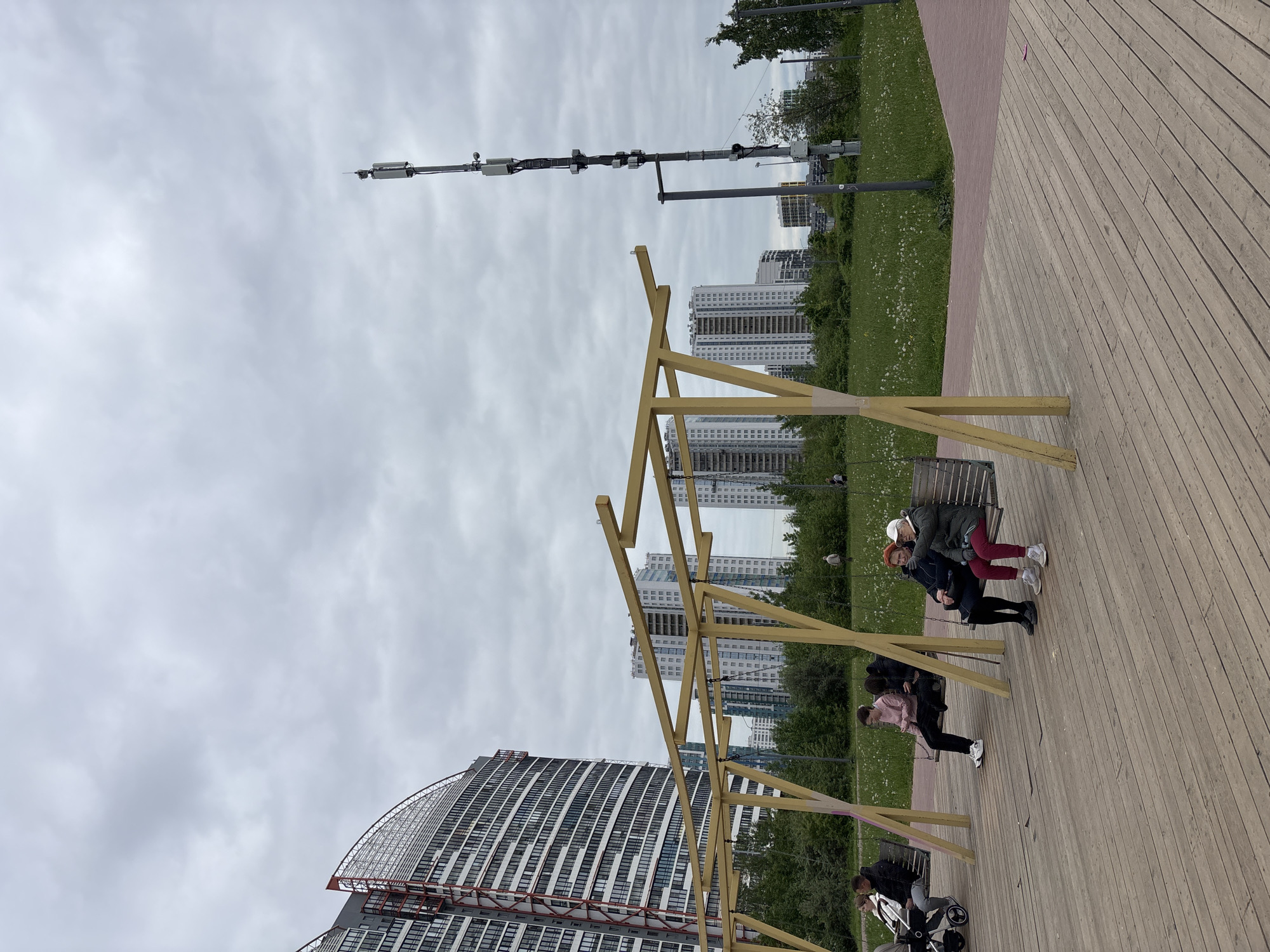 The image depicts a public space, likely a park, in Saint Petersburg, Russia, given the architectural style of the buildings in the background. The foreground features a wooden platform with a light-colored wooden structure. People are present: some are sitting on benches, others are standing nearby. The sky is overcast, and the weather appears mild. A tall pole with equipment, possibly telecommunications hardware, is visible in the middle ground, further back from the public spaces, along with high-rise buildings and a grassy area with trees. There is a tall building in the bottom left part of the image, with a very obvious architectural design, that makes it distinctive.