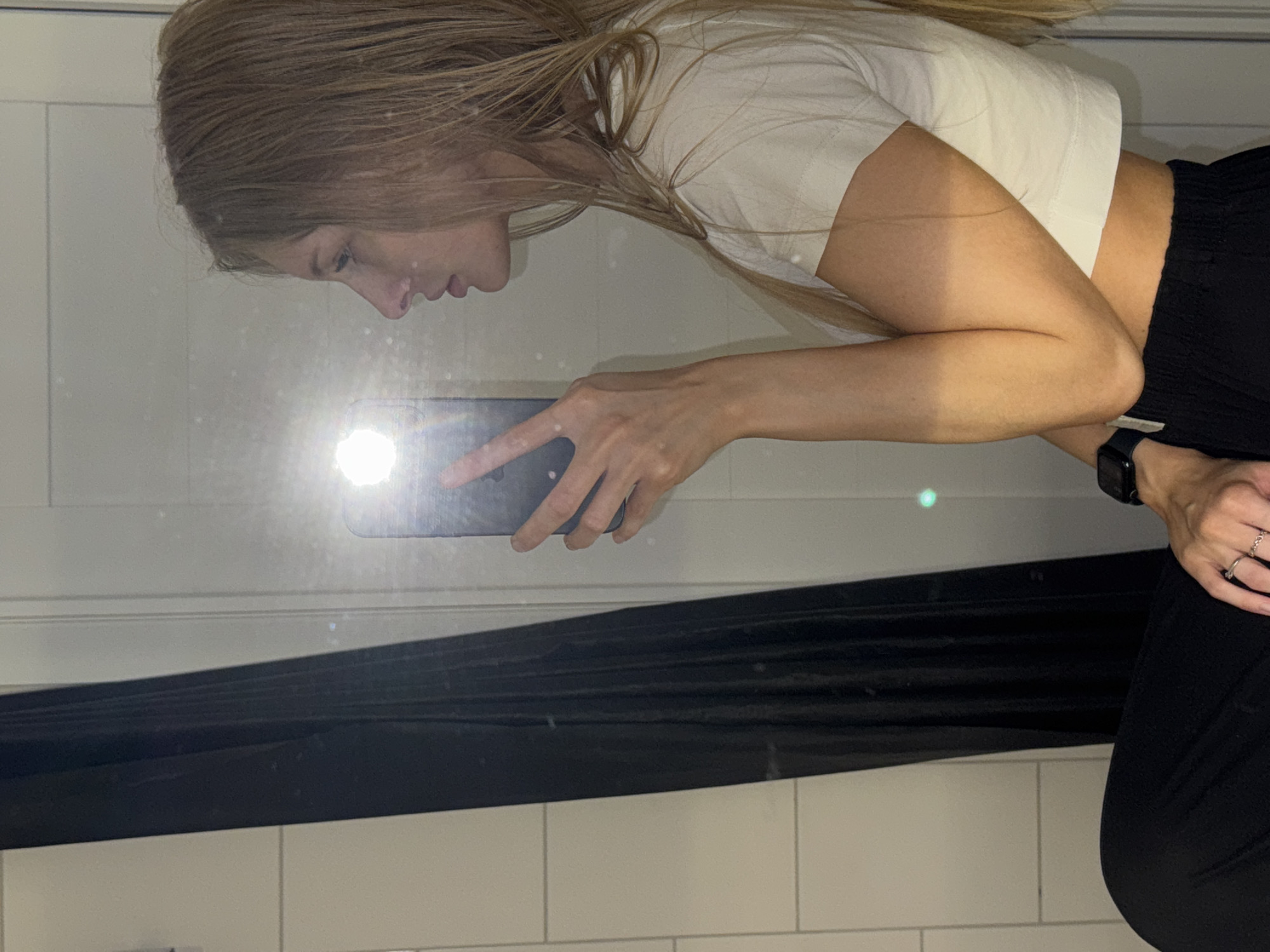 A young Caucasian woman with long blonde hair is shown in a bathroom, taking a selfie in a mirror. She is wearing a white cropped t-shirt and black pants. Her left arm is raised, holding a smartphone. A black shower curtain is visible to her left, and the bathroom walls are tiled. The scene is lit by a bright flash from the phone, which is reflected in the mirror and creates a lens flare. The overall impression is a casual, personal moment captured in a private setting.